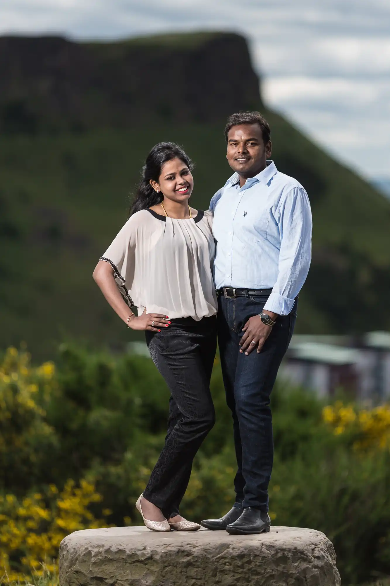 A woman and a man stand side by side on a large rock. They are outdoors with green hills and a clear sky in the background. The man has his arm around the woman's shoulder.