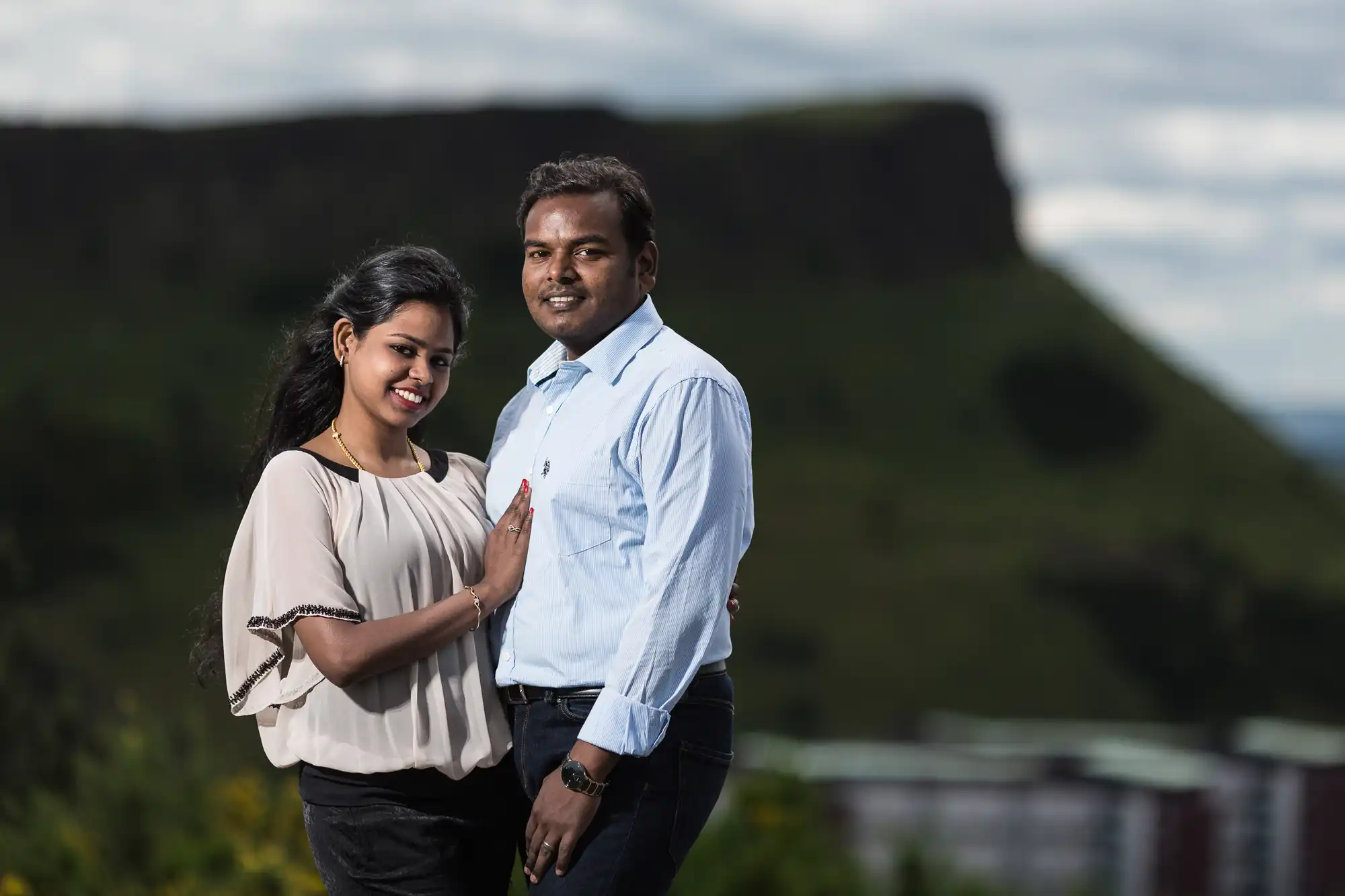 A man and a woman are standing together outdoors with a scenic mountainous background. They are both smiling, and the woman is touching the man's chest while posing for the photo.