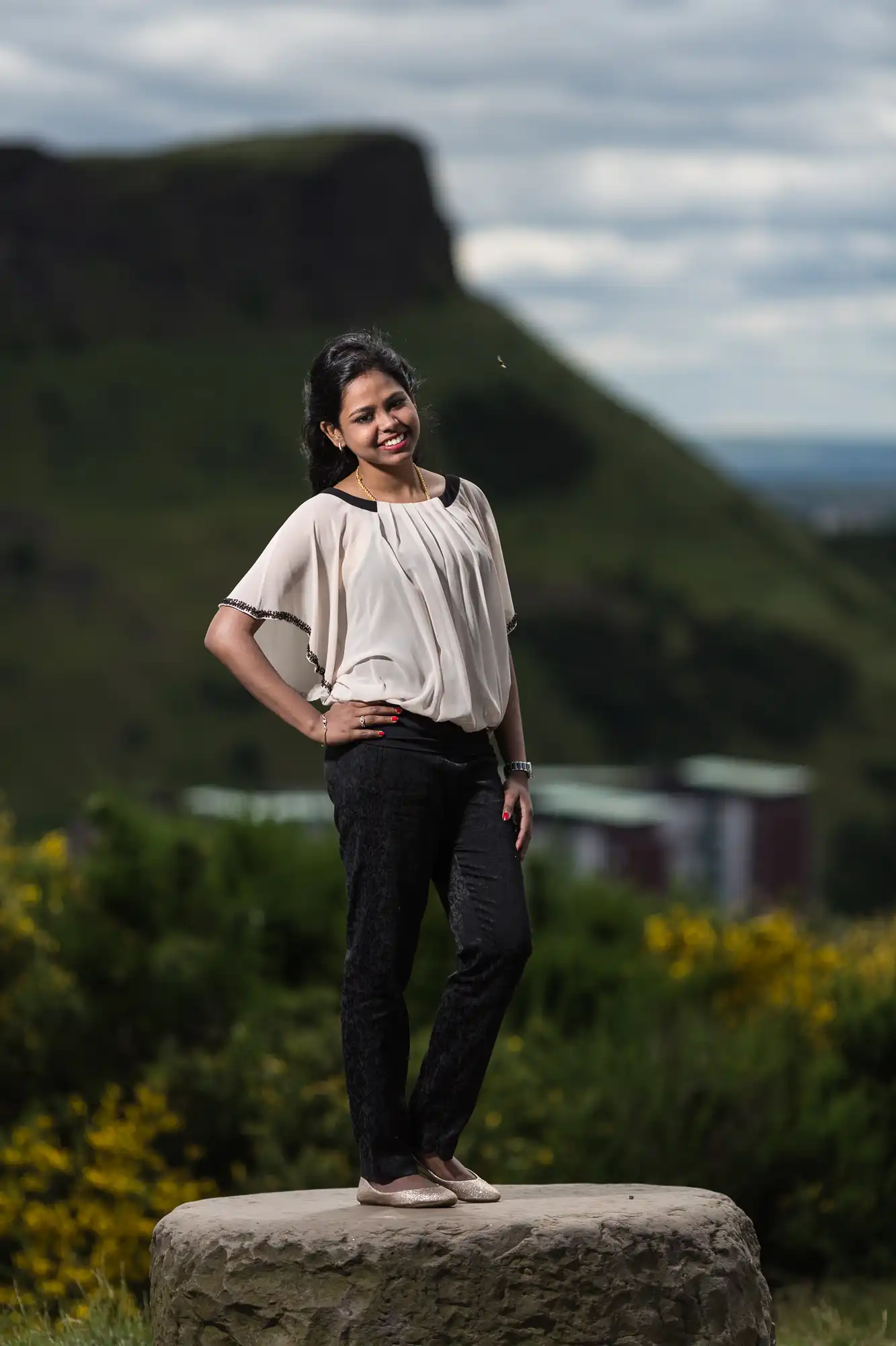 A woman stands on a rock wearing a white blouse and black pants, posing outdoors with hills and greenery in the background.