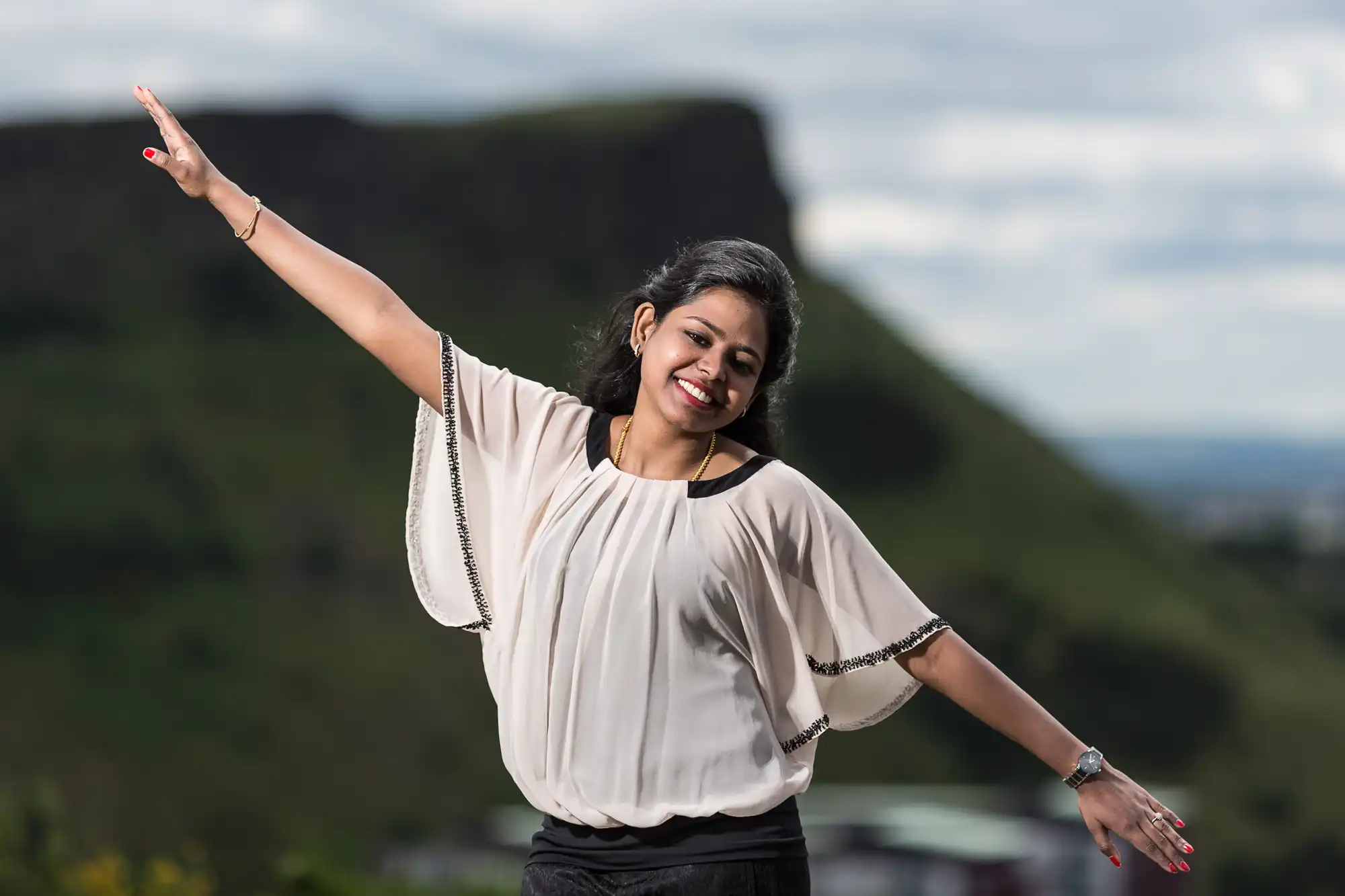 Woman with arms outstretched, smiling, standing outdoors with a blurred mountain and cloudy sky in the background.