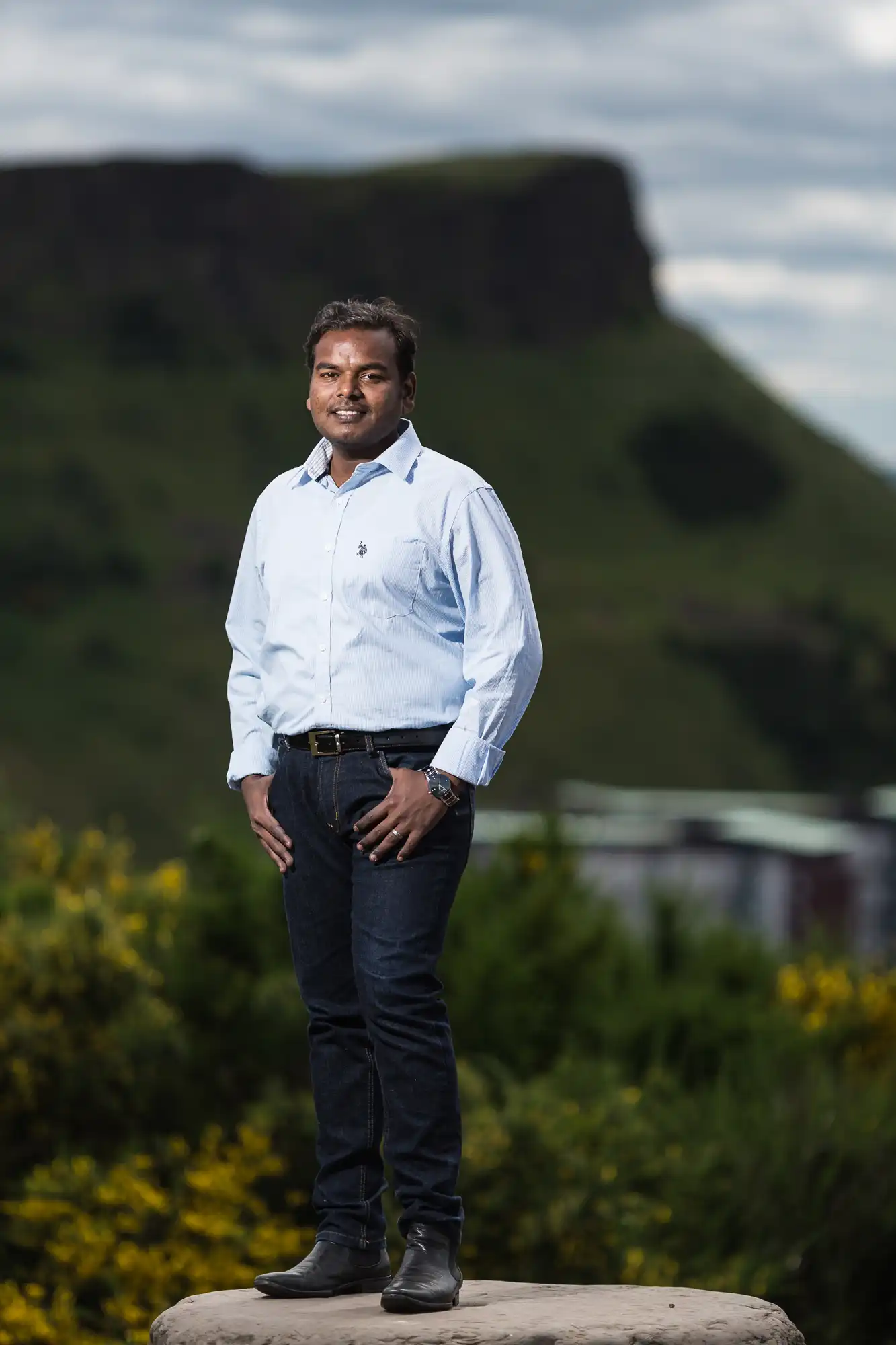 A man in a light blue shirt and dark jeans stands confidently outdoors with grassy hills and a cliff in the background.