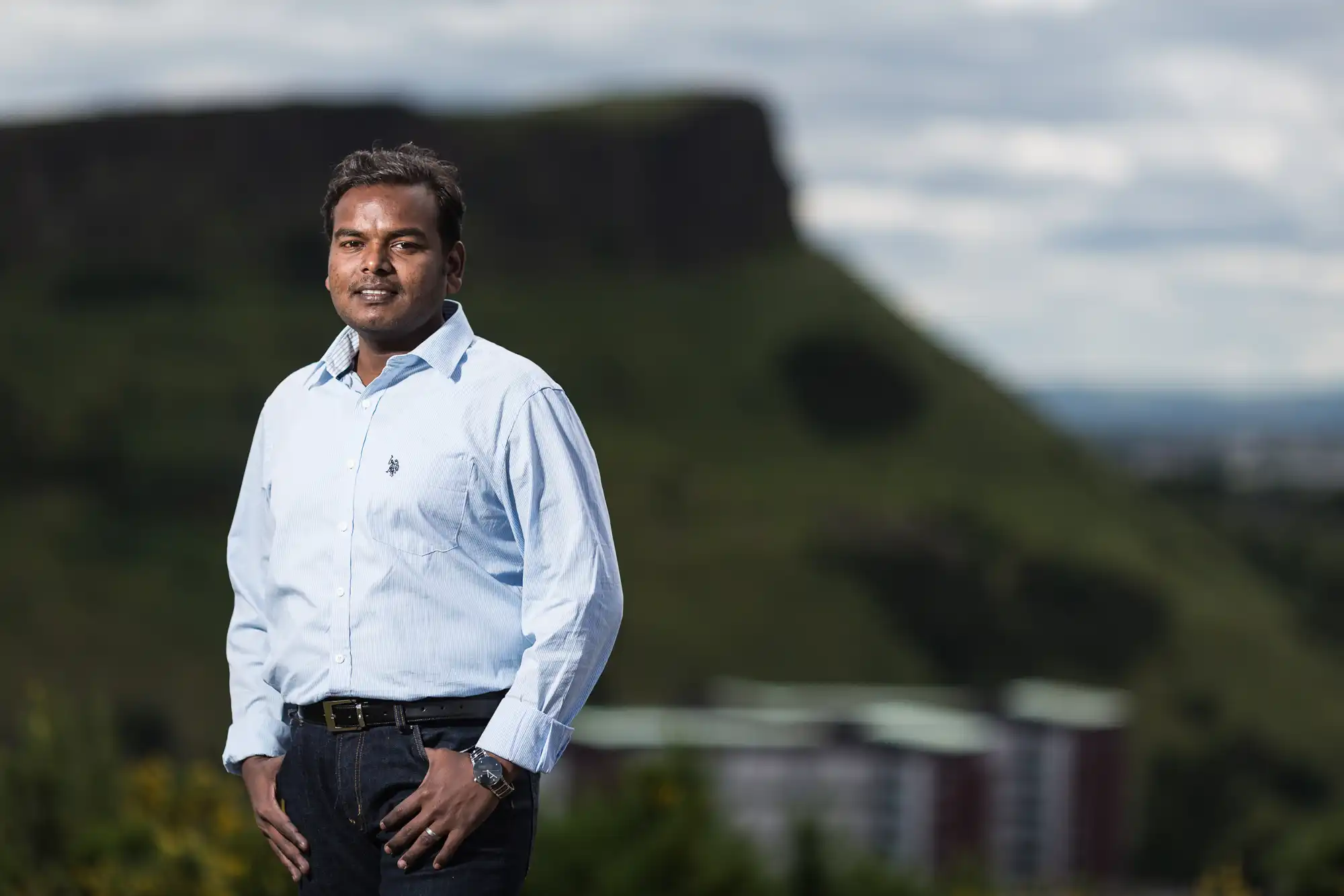 A man in a light blue shirt and jeans stands outdoors against a blurred backdrop of hills and buildings, looking at the camera.