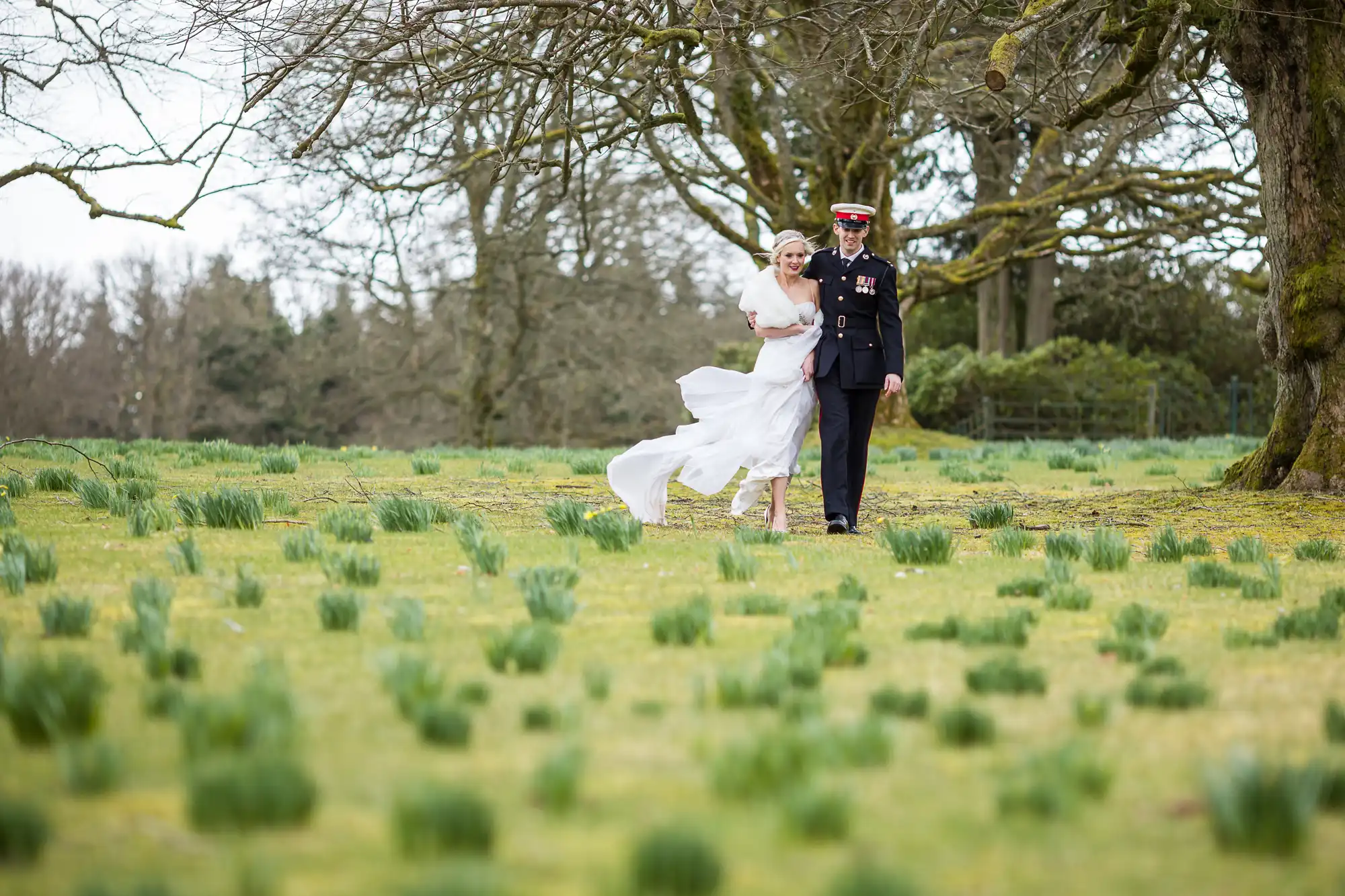 A bride and a uniformed groom walk through a grassy field with scattered plants and bare trees in the background. The bride's dress flows in the wind.