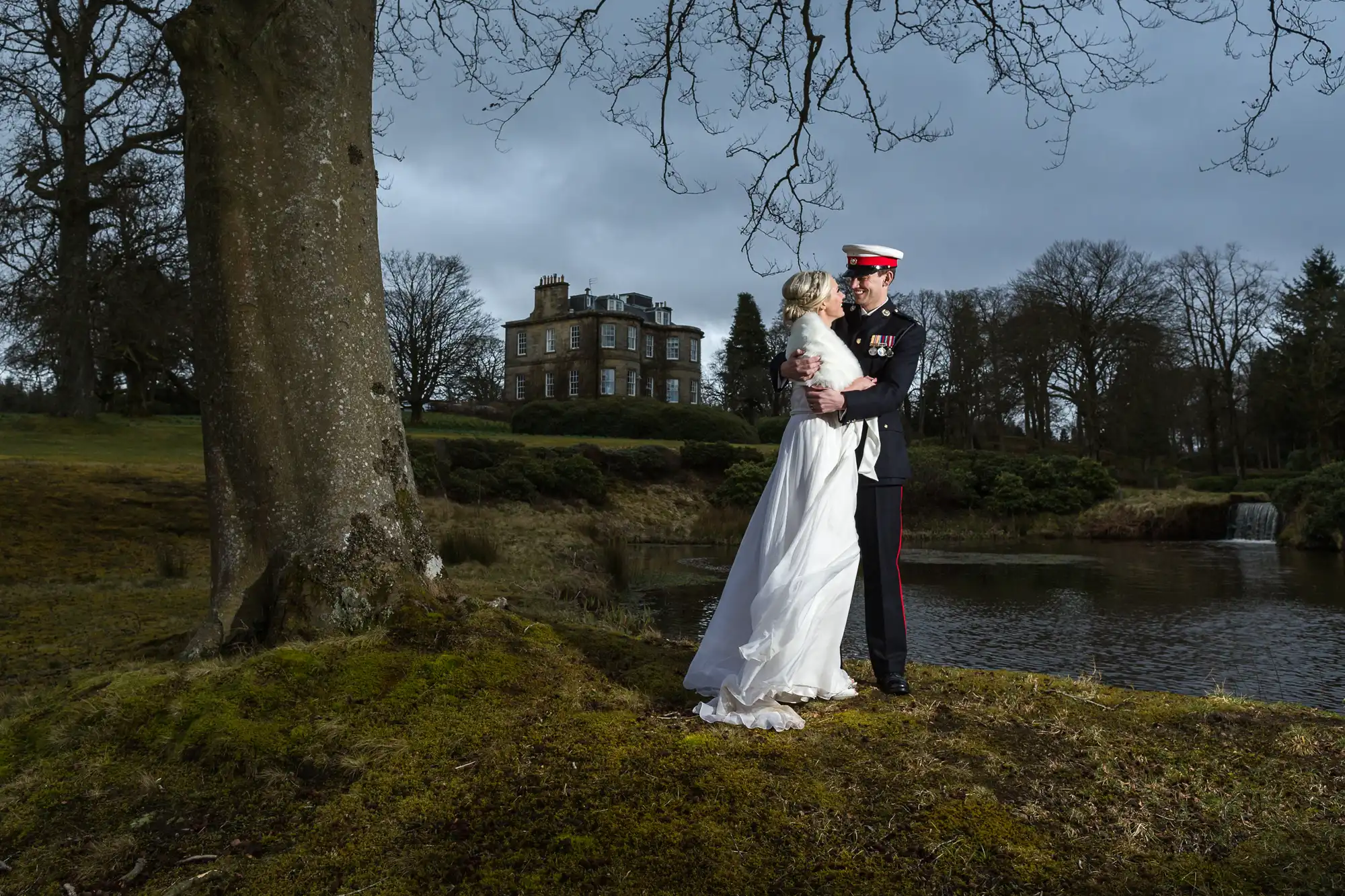 Bride in a white dress and groom in a military uniform embracing under a tree by a pond, with a historic building and bare trees in the background.