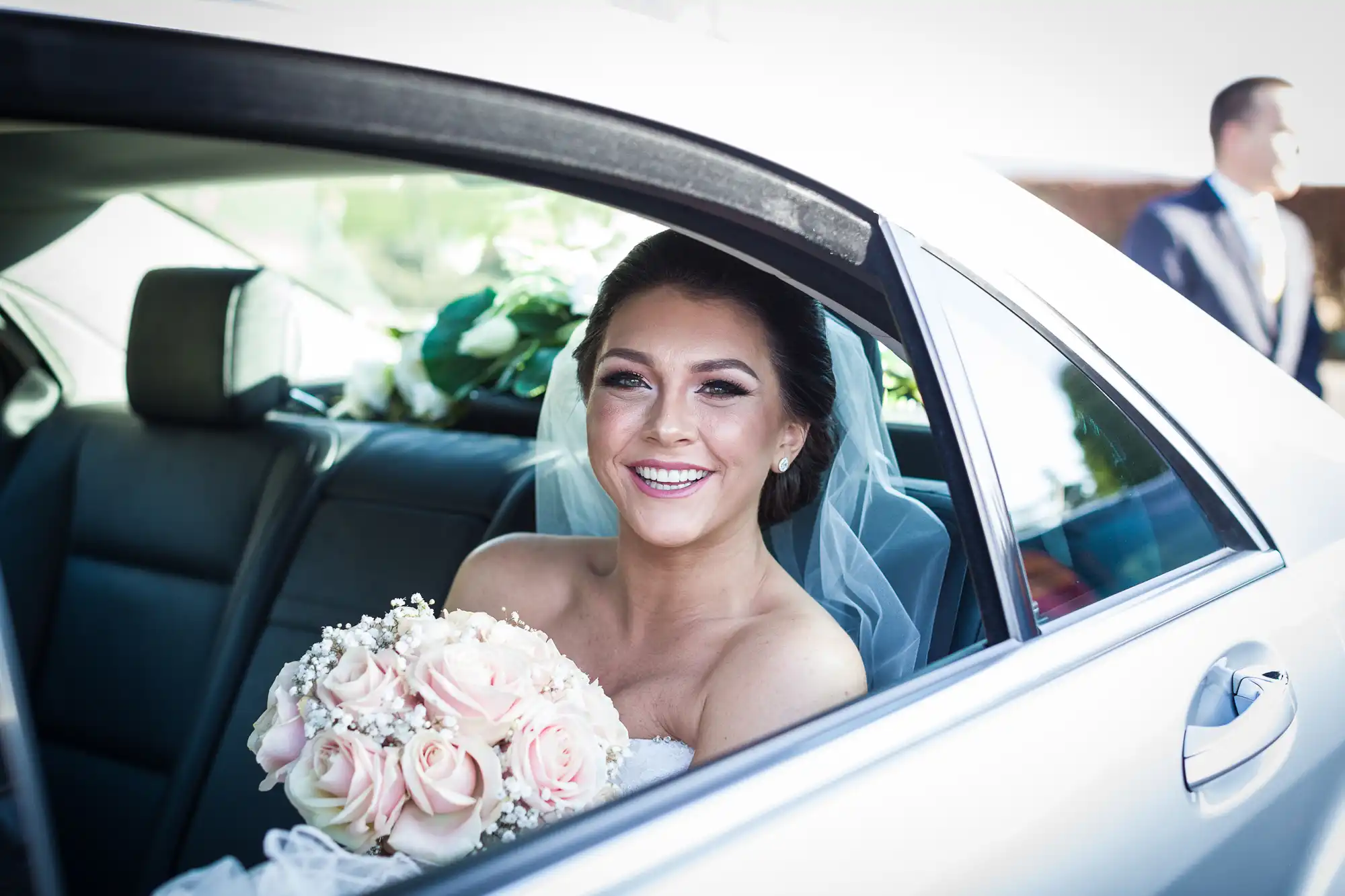 A bride sits in the backseat of a car, smiling and holding a bouquet of pink roses with white accents.