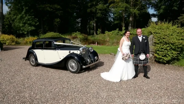 The Barn at Harburn wedding video: A bride and groom pose for a photo beside a vintage black and white car on a gravel path, with greenery and trees in the background.