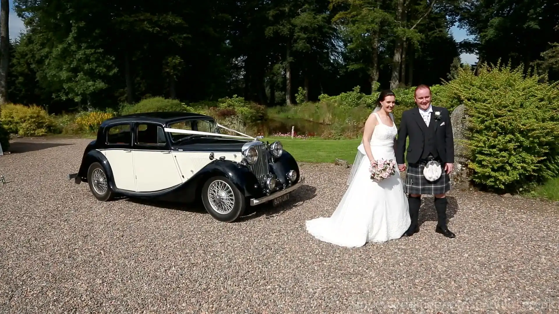 The Barn at Harburn wedding video: A bride and groom pose for a photo beside a vintage black and white car on a gravel path, with greenery and trees in the background.