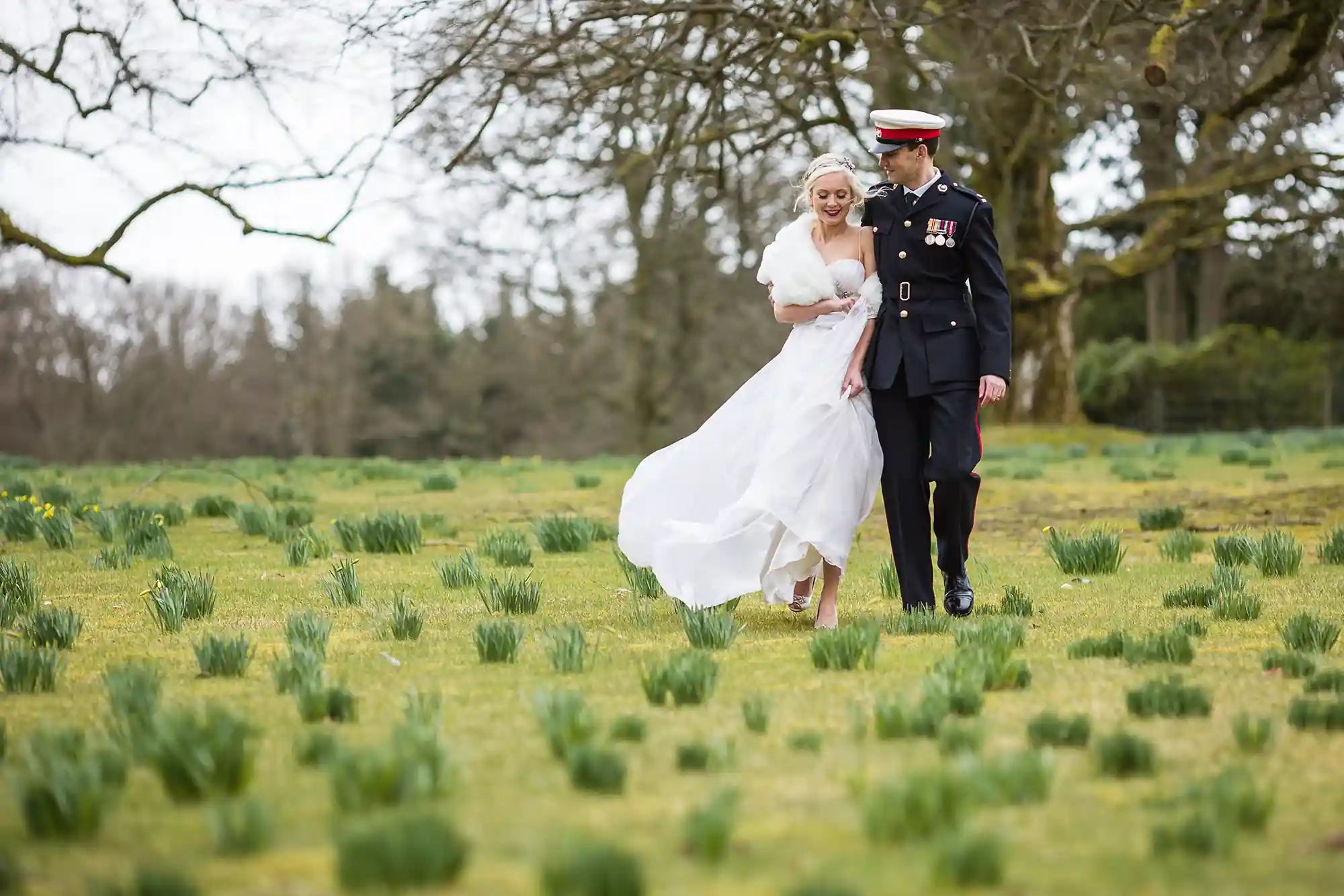 A bride in a white dress and a groom in military uniform walking hand-in-hand through a field with young green plants, with trees in the background.