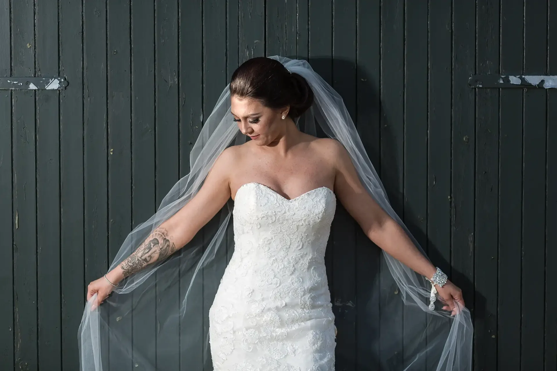 A bride in a white strapless gown adjusts her veil against a dark wooden wall, sunlight highlighting her and the textures of her dress.