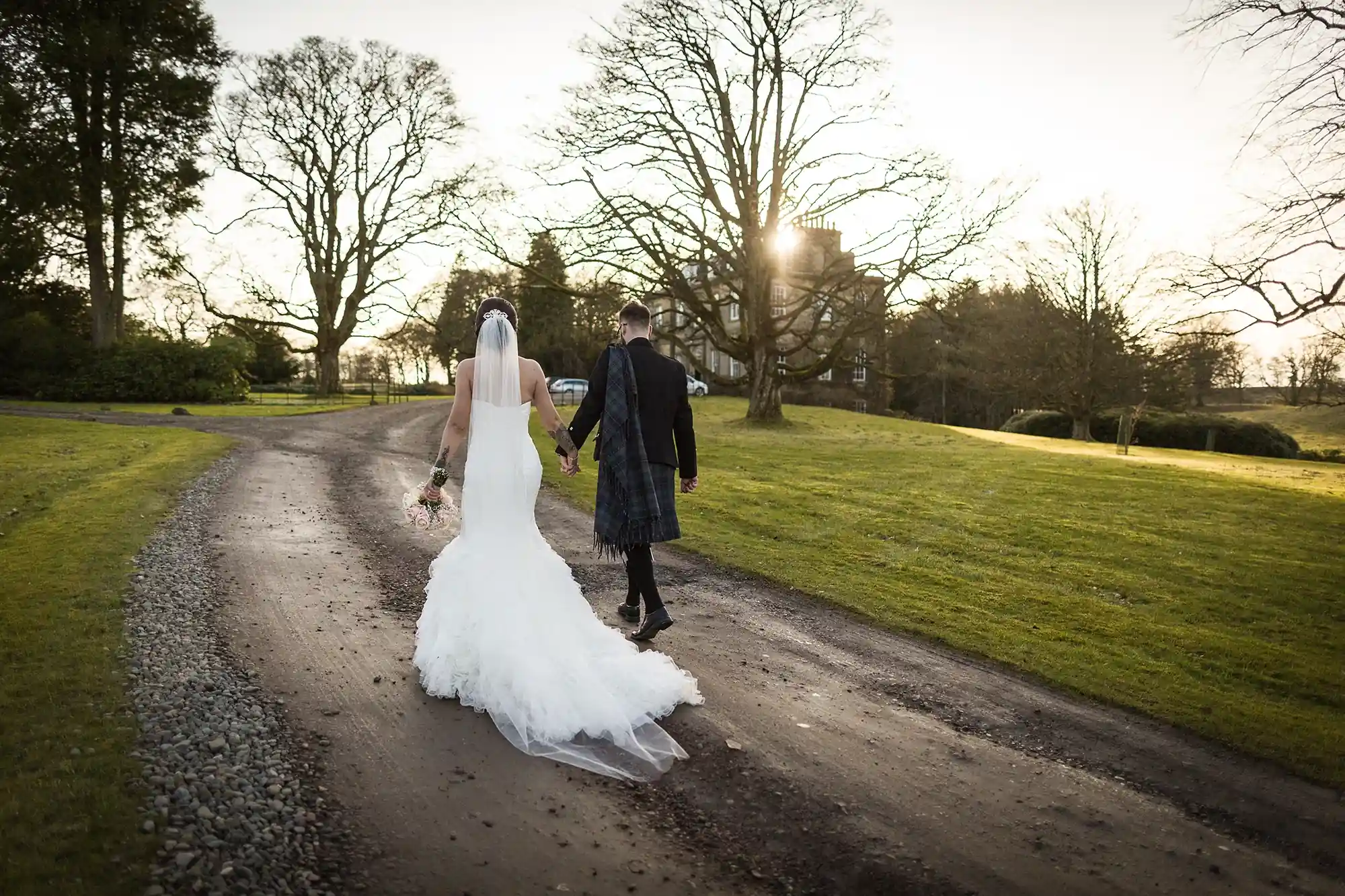 A bride and groom holding hands while walking down a tree-lined pathway at sunset.