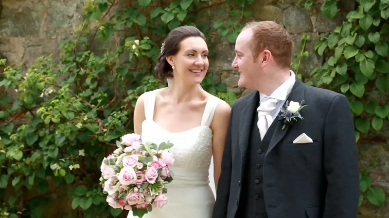 The Barn at Harburn wedding video: A bride and groom smile at each other. The bride holds a bouquet of pink roses. They stand in front of foliage-covered stone.