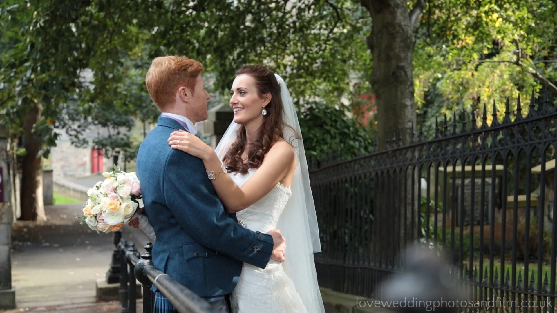 The Caledonian Edinburgh wedding video image of newlyweds Joy and Nick, a bride and groom stand close together outdoors, smiling at each other near a black iron fence, with trees and greenery in the background.