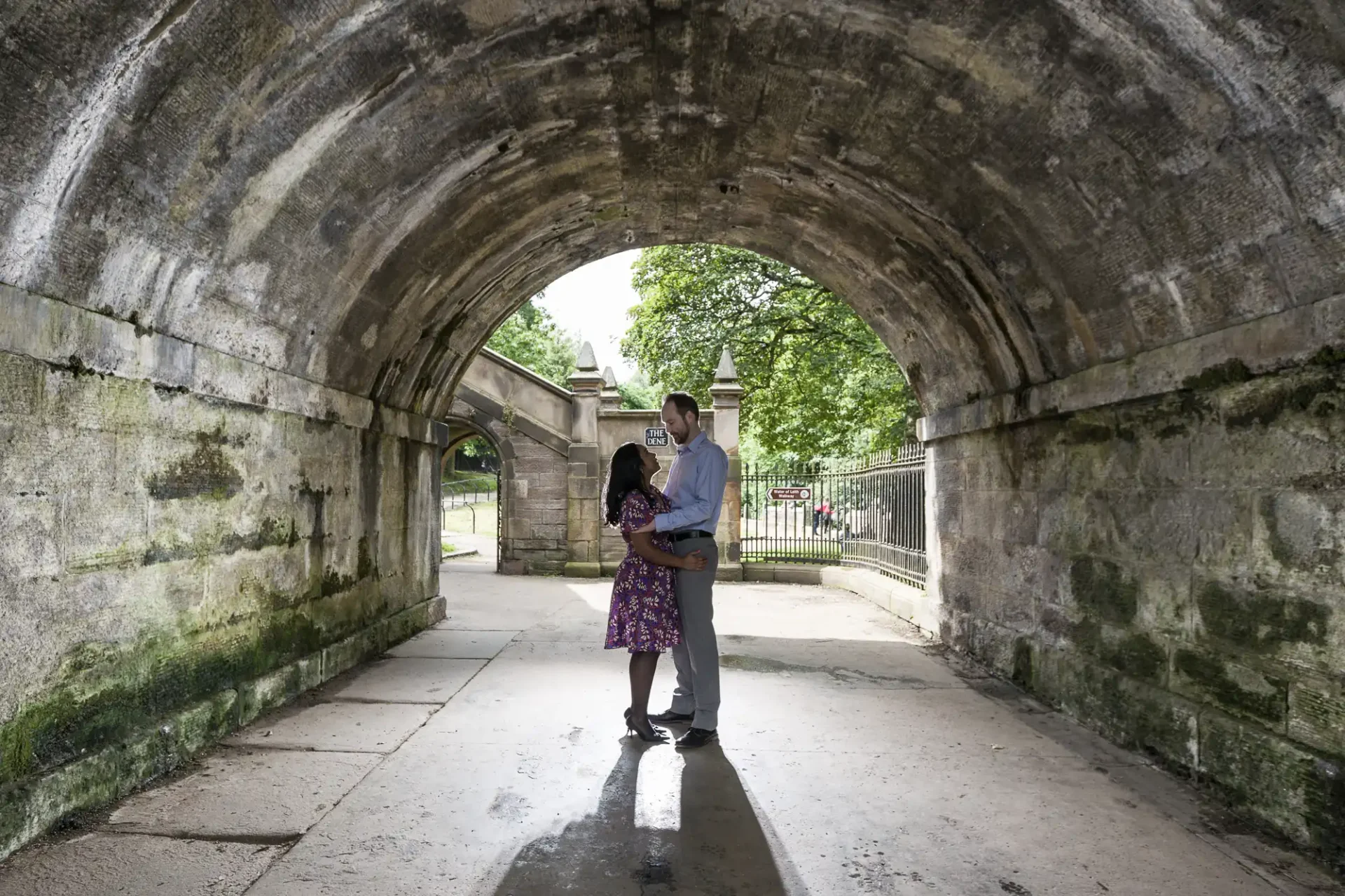 A couple stands facing each other under a stone archway in an outdoor setting with trees and a gate visible in the background.