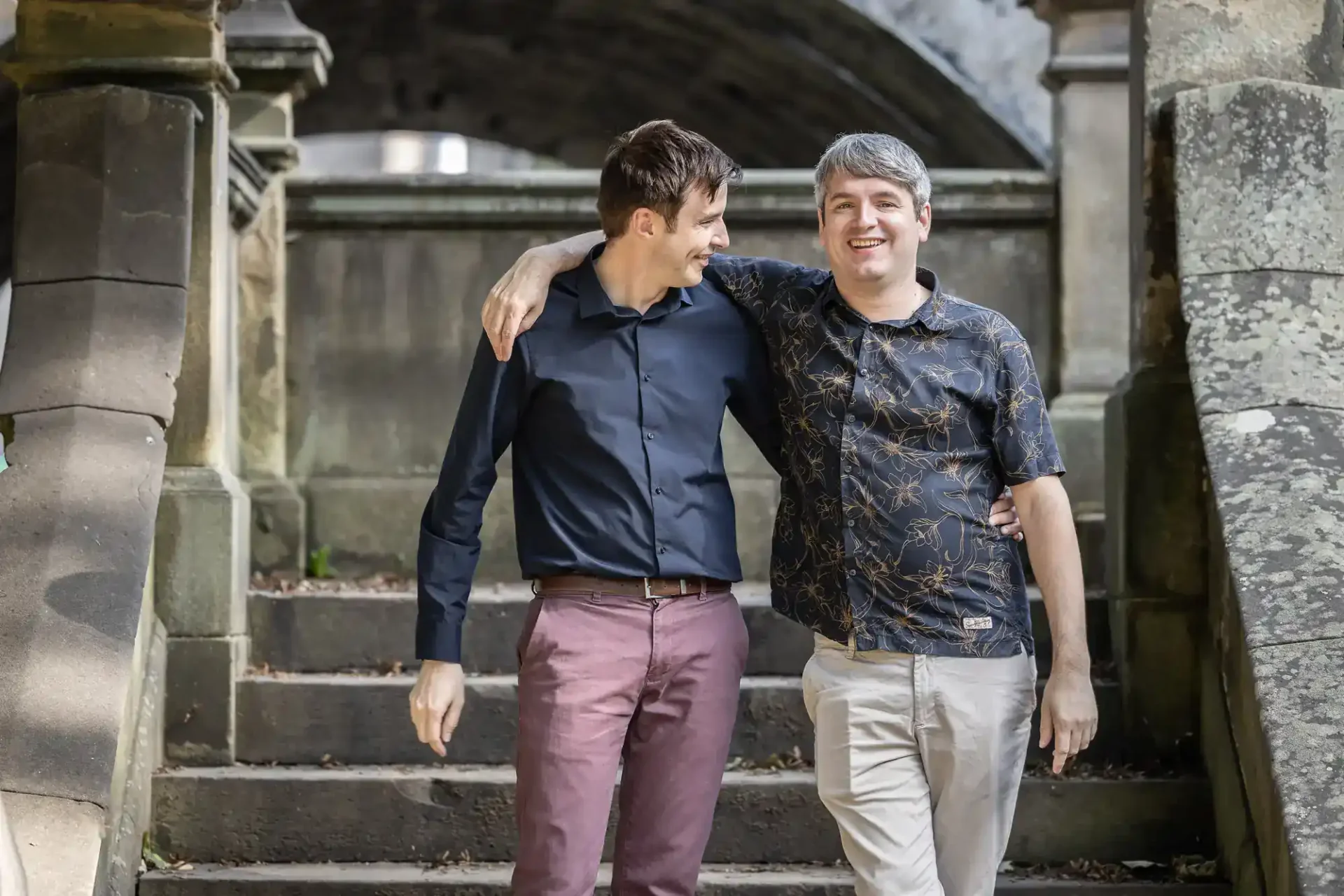 Two men walking down stone steps outside, smiling with their arms around each other's shoulders.