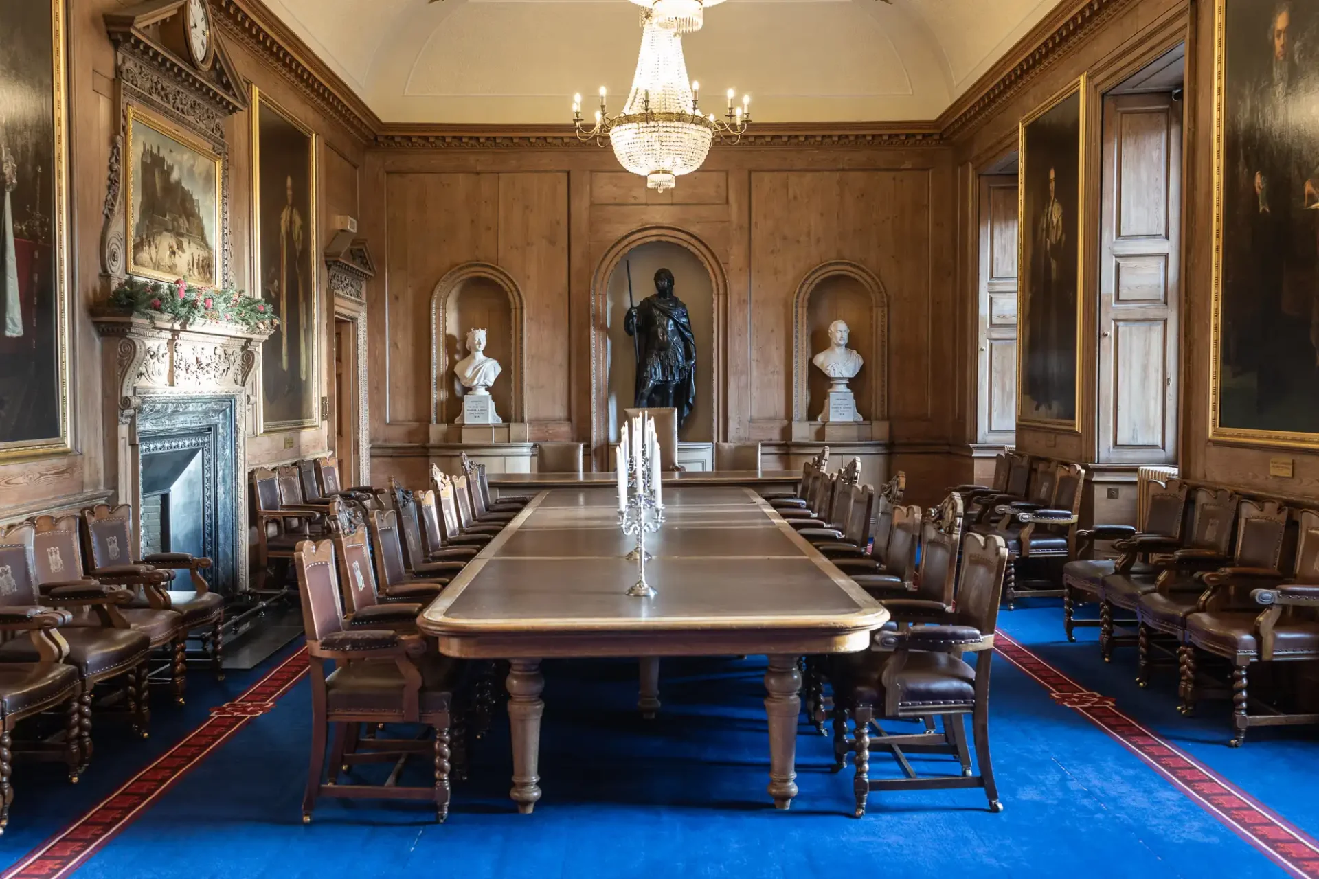 A formal dining room with a long wooden table, surrounded by chairs. Portraits adorn the wood-paneled walls, and a chandelier hangs from the ceiling. Busts are displayed in the background.