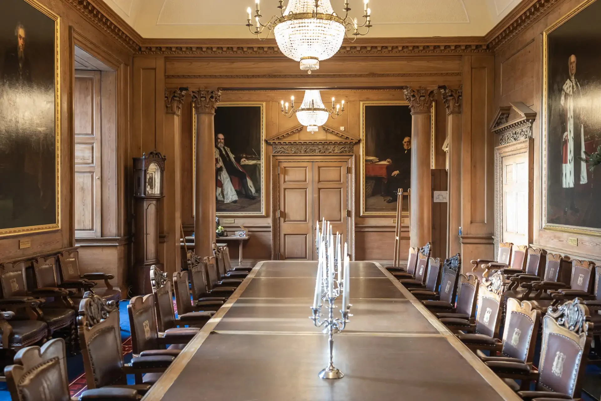 Opulent wood-paneled dining room with a long table, surrounded by ornate chairs, chandeliers above, and large portraits on the walls.