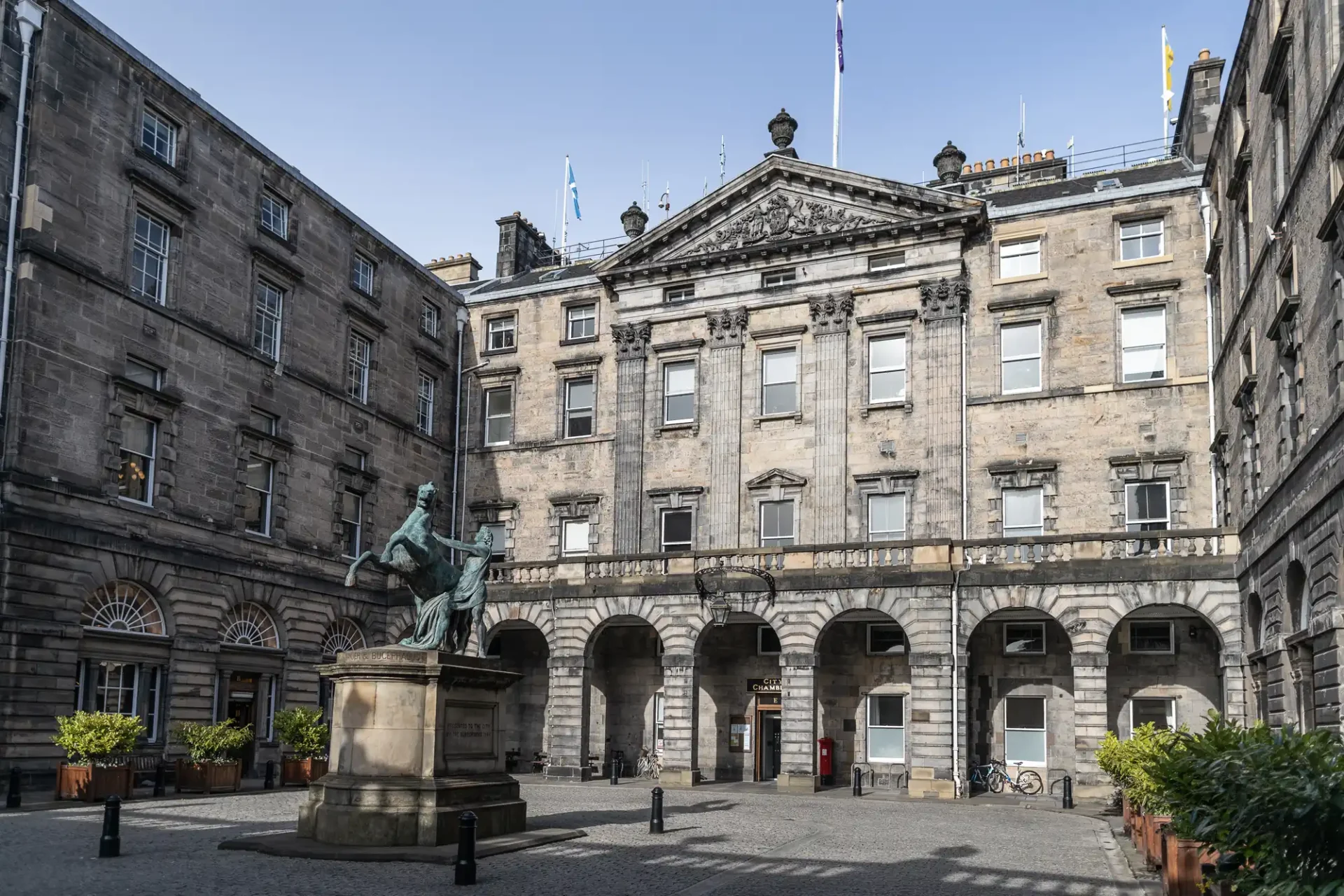 A historic stone building with arched windows surrounds a courtyard featuring a statue of a seated figure on horseback. The sky is clear, and potted plants line the courtyard's edges.