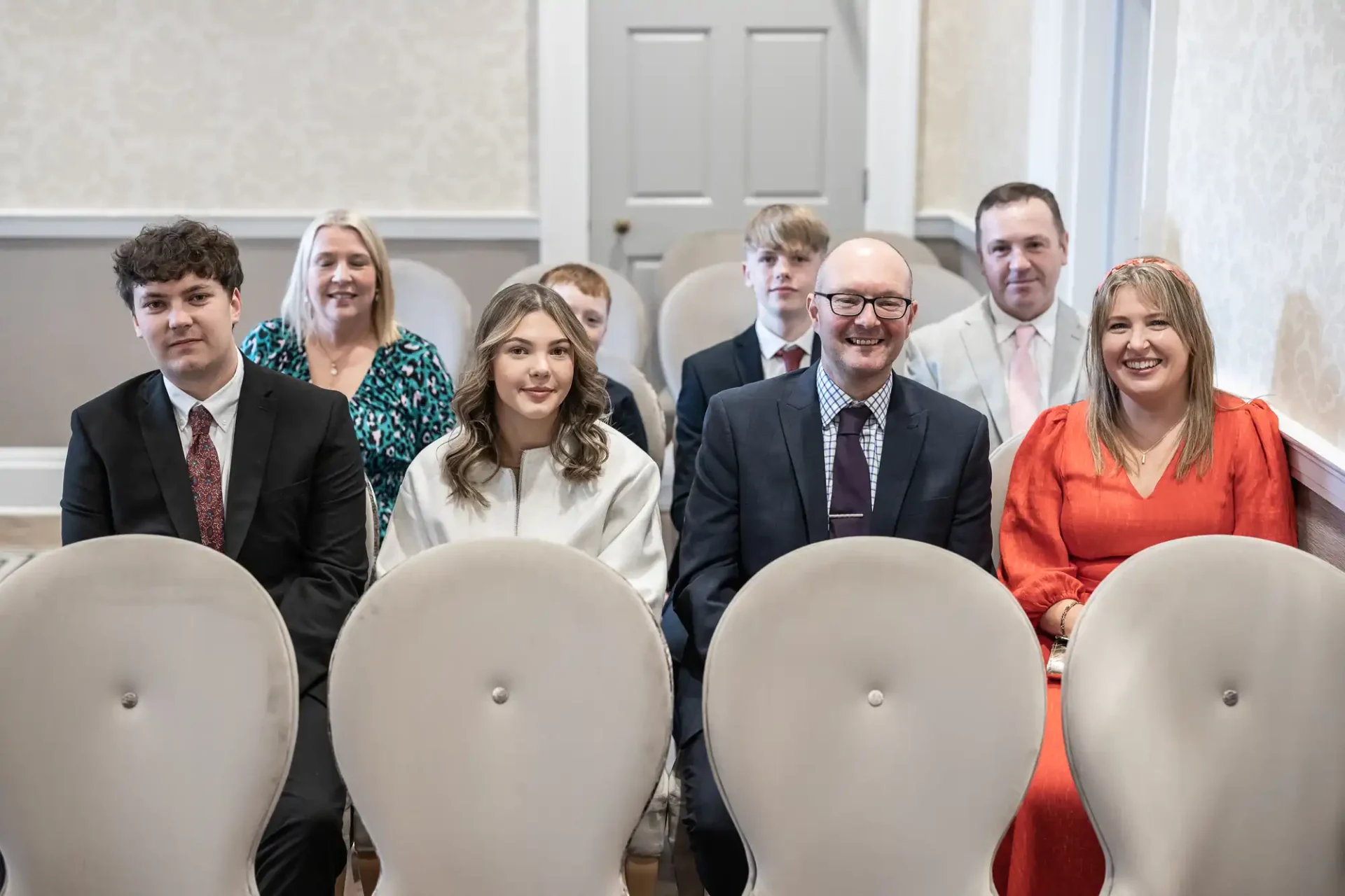 Group of people sitting, dressed formally, in a room with beige walls.