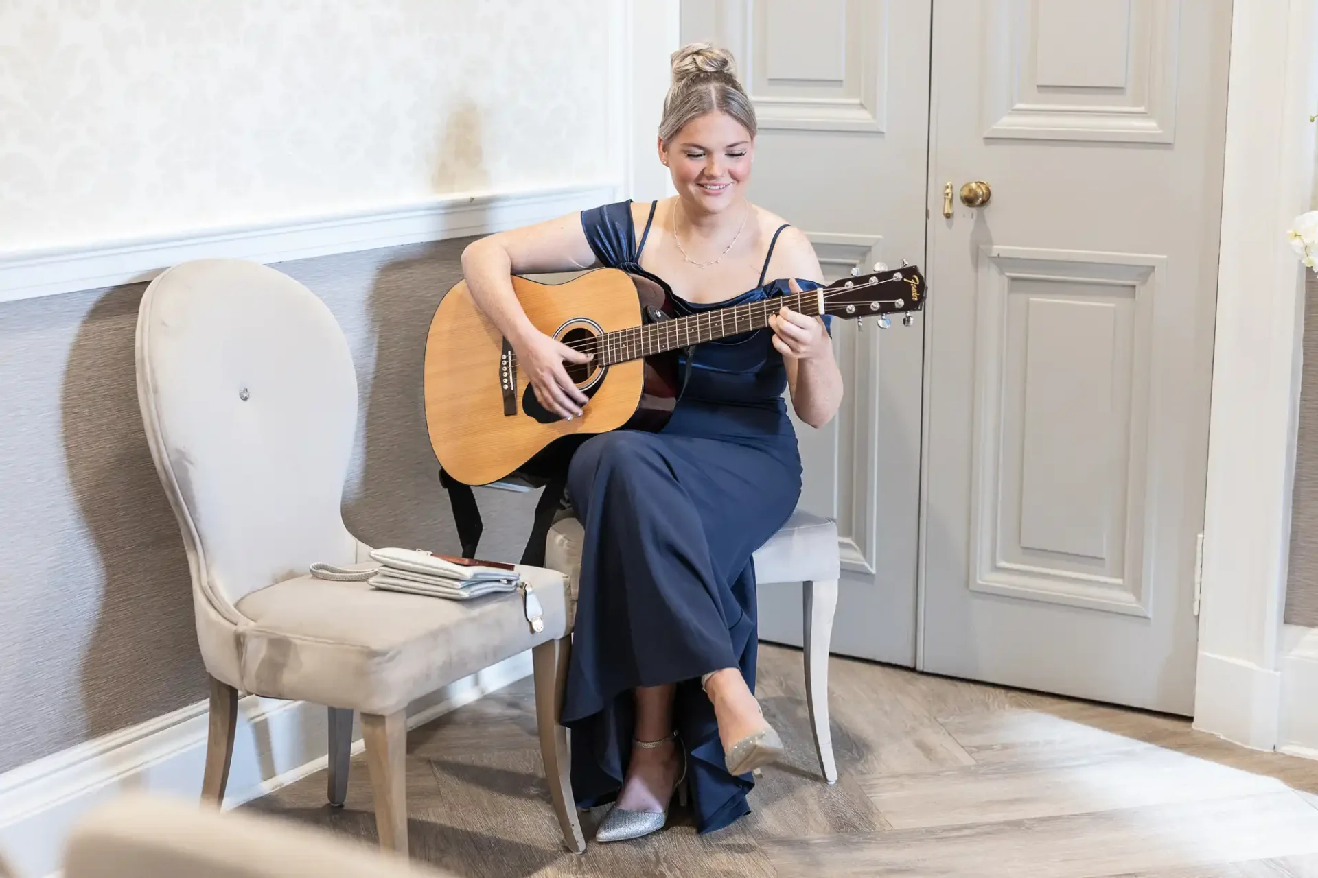 A woman in a blue dress sits on a chair, playing an acoustic guitar in a room with light gray walls and a gray door.