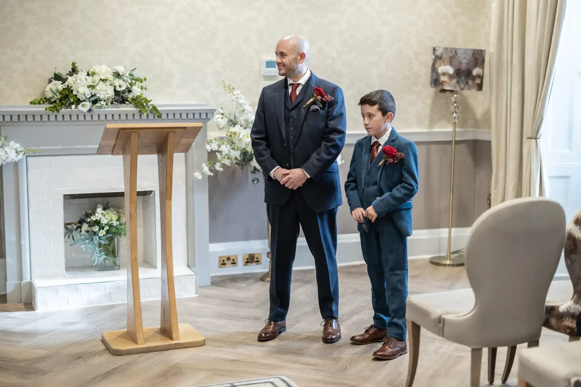 Two people in suits stand by a lectern in a decorated room with floral arrangements and a fireplace.