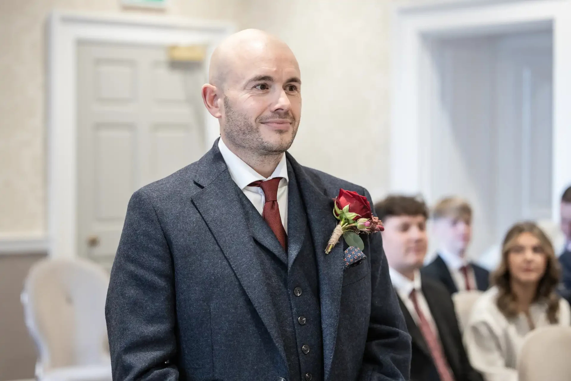 Bald man in a suit with a red rose boutonnière standing indoors, smiling. Seated people in the background.