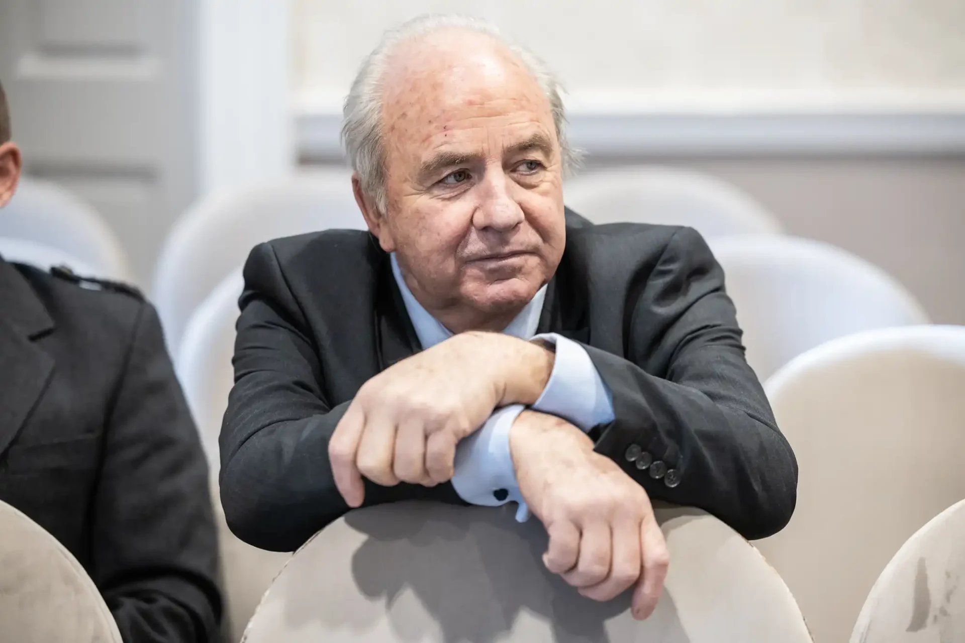 An older man in a suit rests his arms on the back of a white chair, looking contemplative.