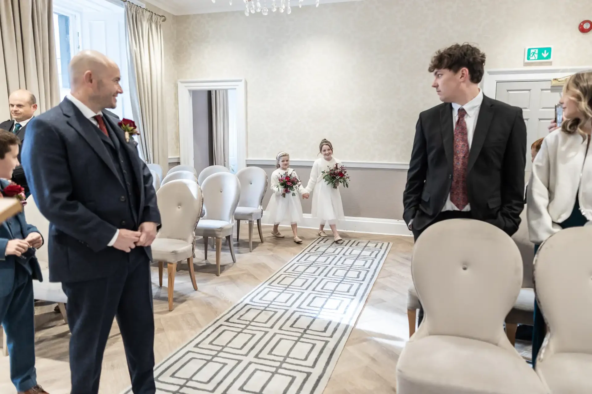 Two flower girls walk down an aisle holding bouquets, approached by a group of people in formal attire waiting at the end. The room is elegantly decorated with beige and white tones.