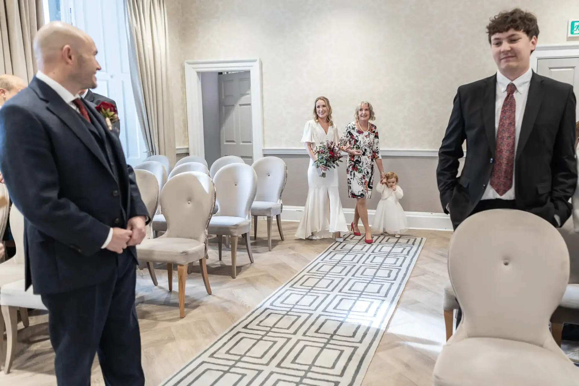 A man in a suit stands at the front as two women, one holding a child, walk down an aisle in a room with empty chairs. Another man in a suit stands nearby.