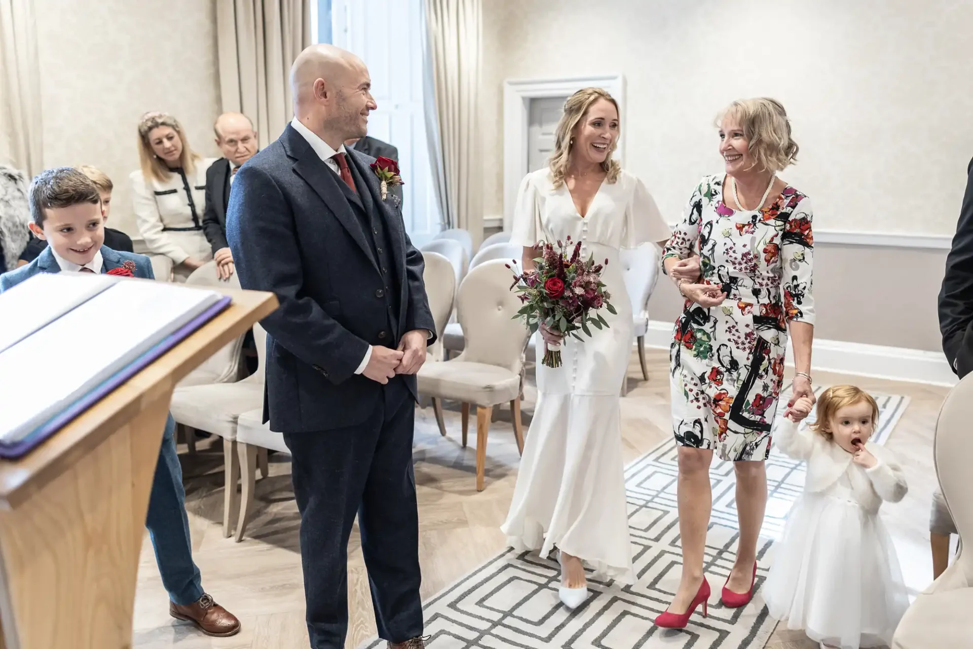 A bride holding a bouquet walks down an aisle with two children and a woman in a floral dress, approaching a smiling man in a suit. Guests are seated in the background.