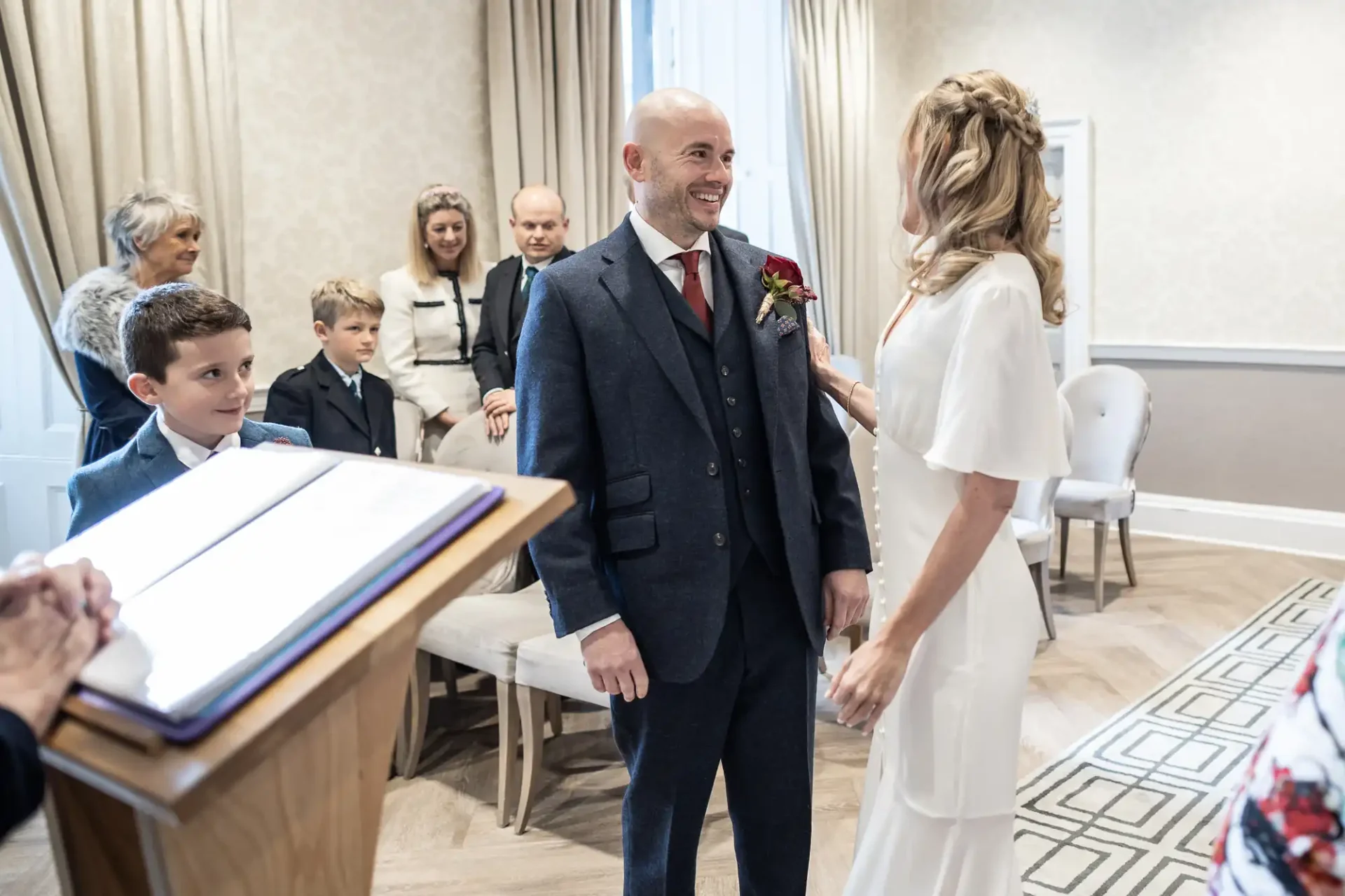 A couple is getting married in a small ceremony room, with guests seated and a child standing nearby.