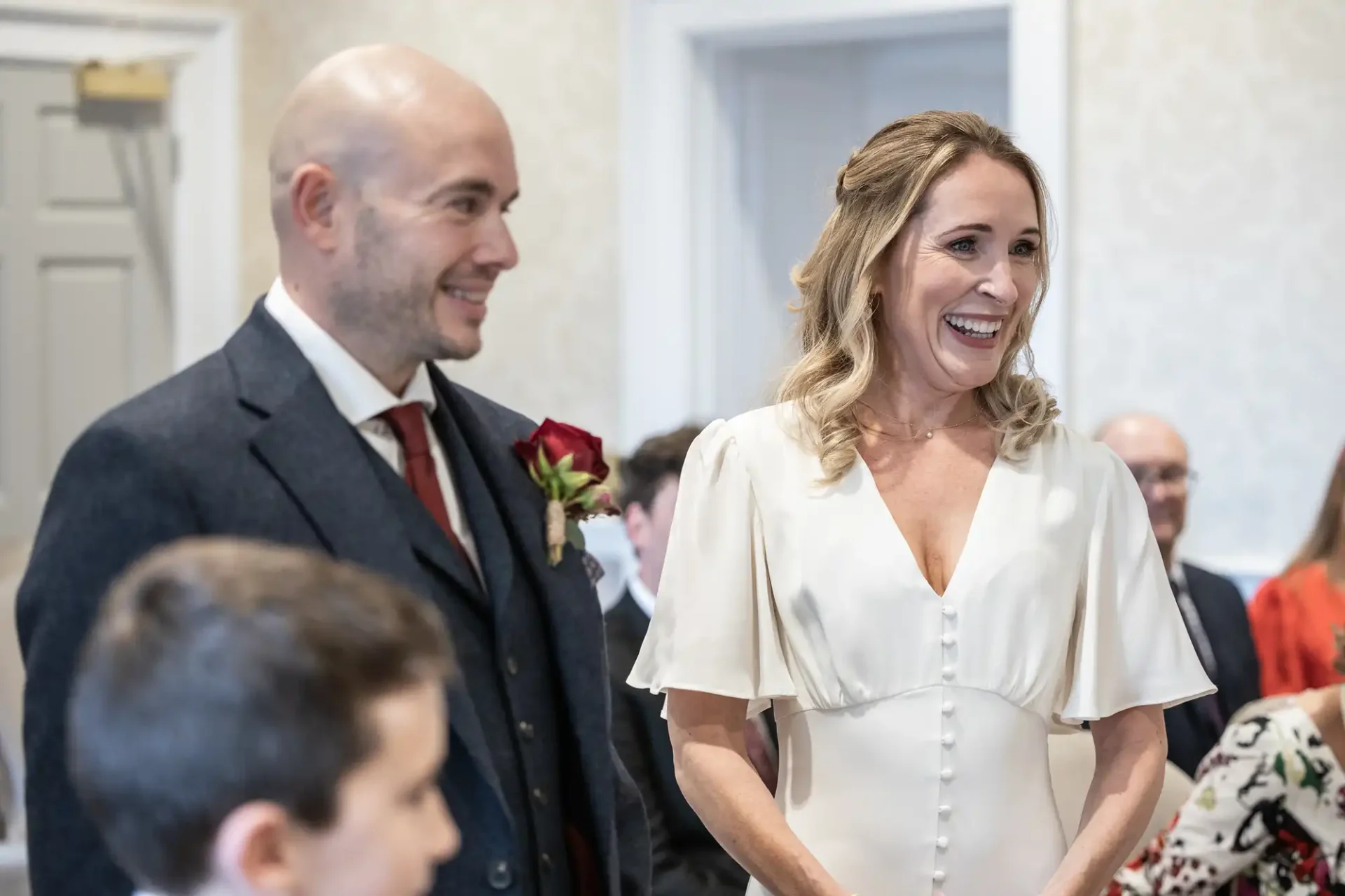 A smiling couple stands indoors at a wedding ceremony, dressed in formal attire. Other guests are seated in the background.