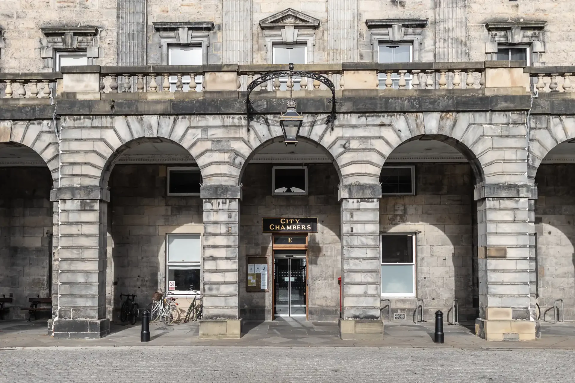 Stone building facade with three arches. The central arch leads to a doorway labeled "City Chambers." Bicycles are parked nearby, and a lamp hangs above the entrance.