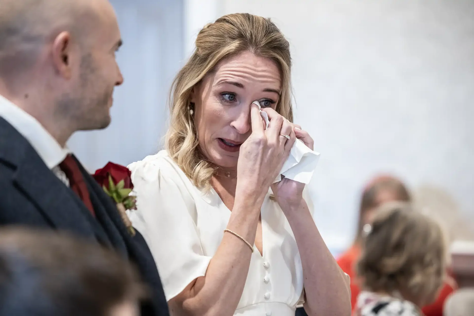 A woman wiping tears with a tissue stands beside a man in a suit, indoors.