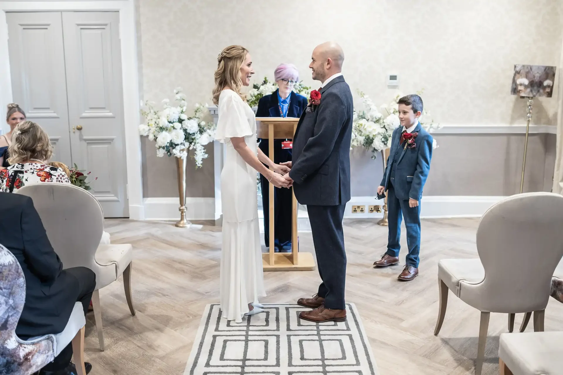 A couple stands holding hands during a wedding ceremony indoors, with a person officiating and a well-dressed child nearby. White flowers decorate the room.