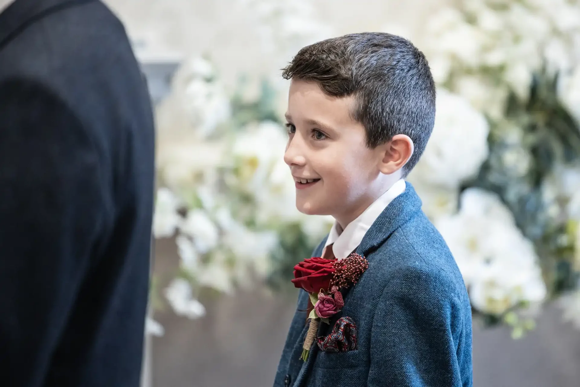 A young boy in a suit and red boutonniere stands smiling, with floral decorations in the background.