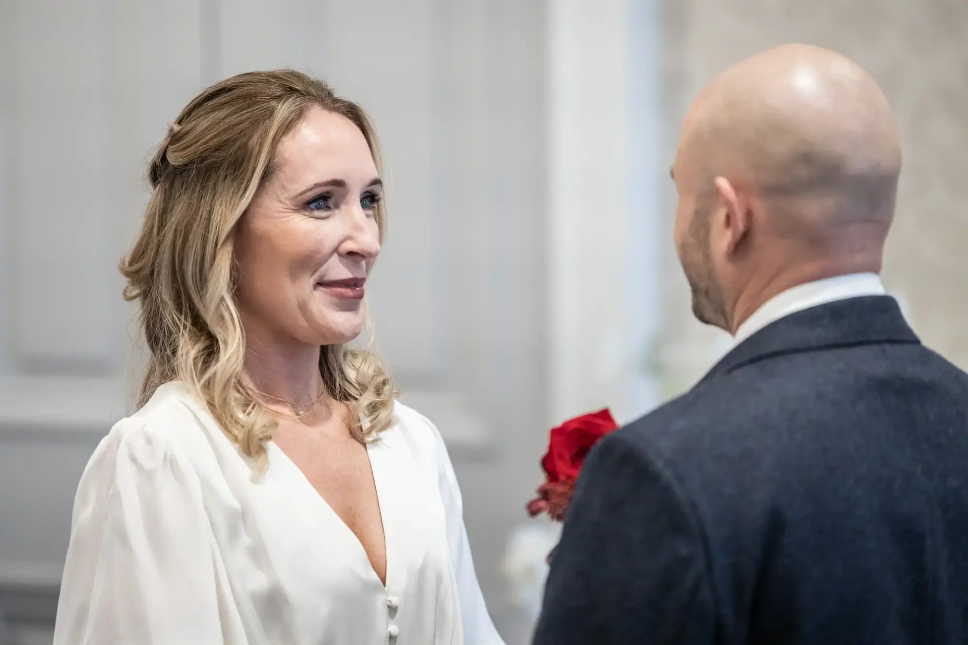 A woman in a white dress and a man in a dark suit stand facing each other indoors. The man holds red flowers.