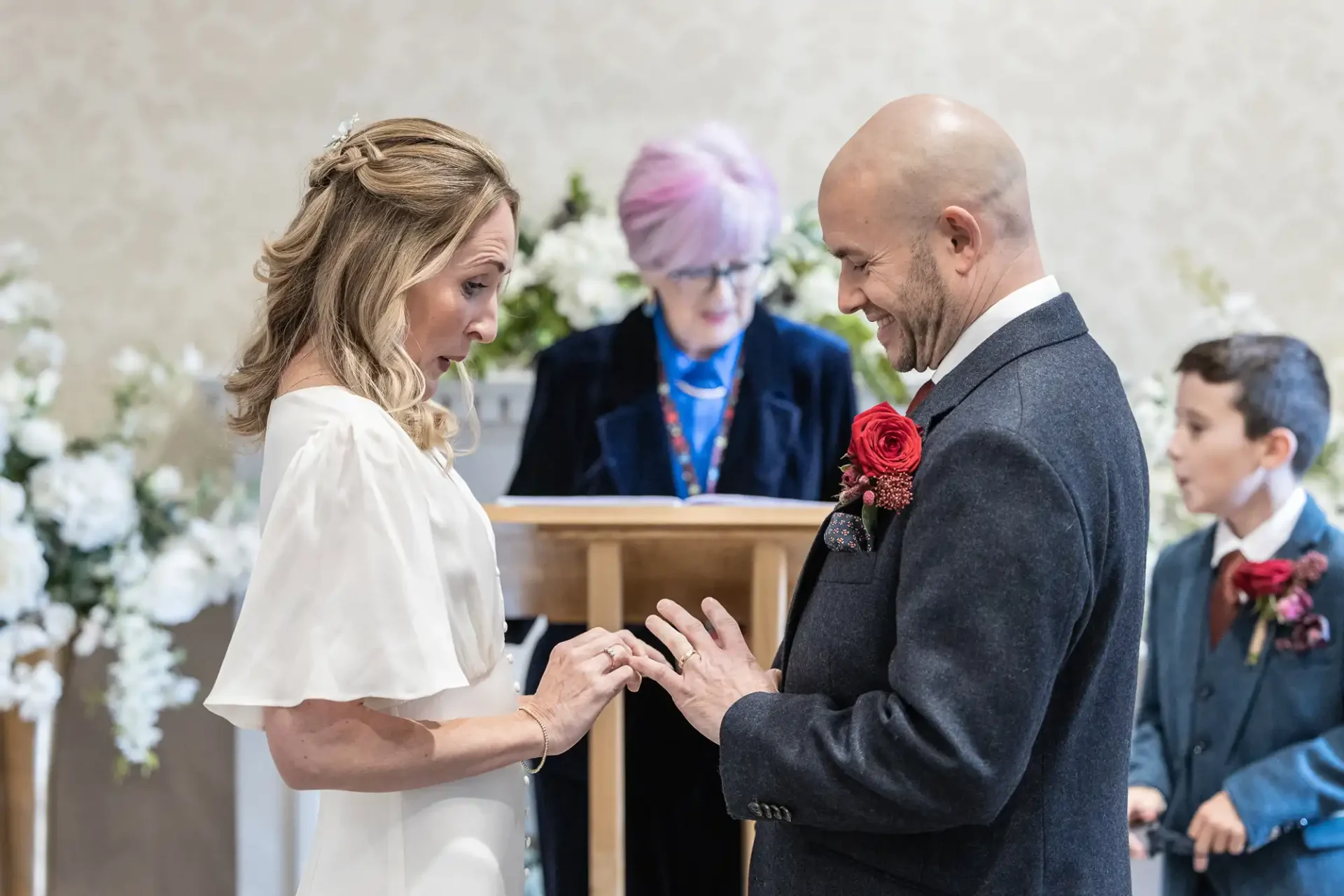 A couple exchanges rings during a wedding ceremony, with a celebrant and a child in the background.