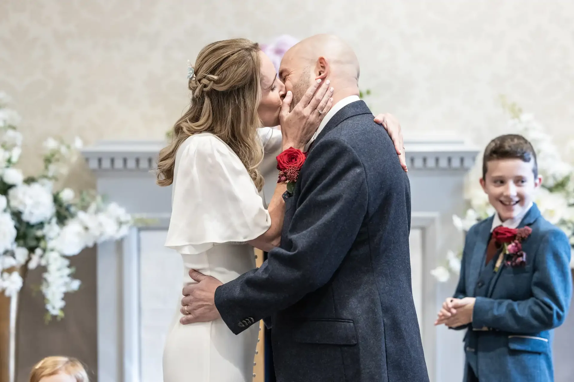 A couple kisses at their wedding ceremony, surrounded by white flowers. A child in a suit smiles beside them.