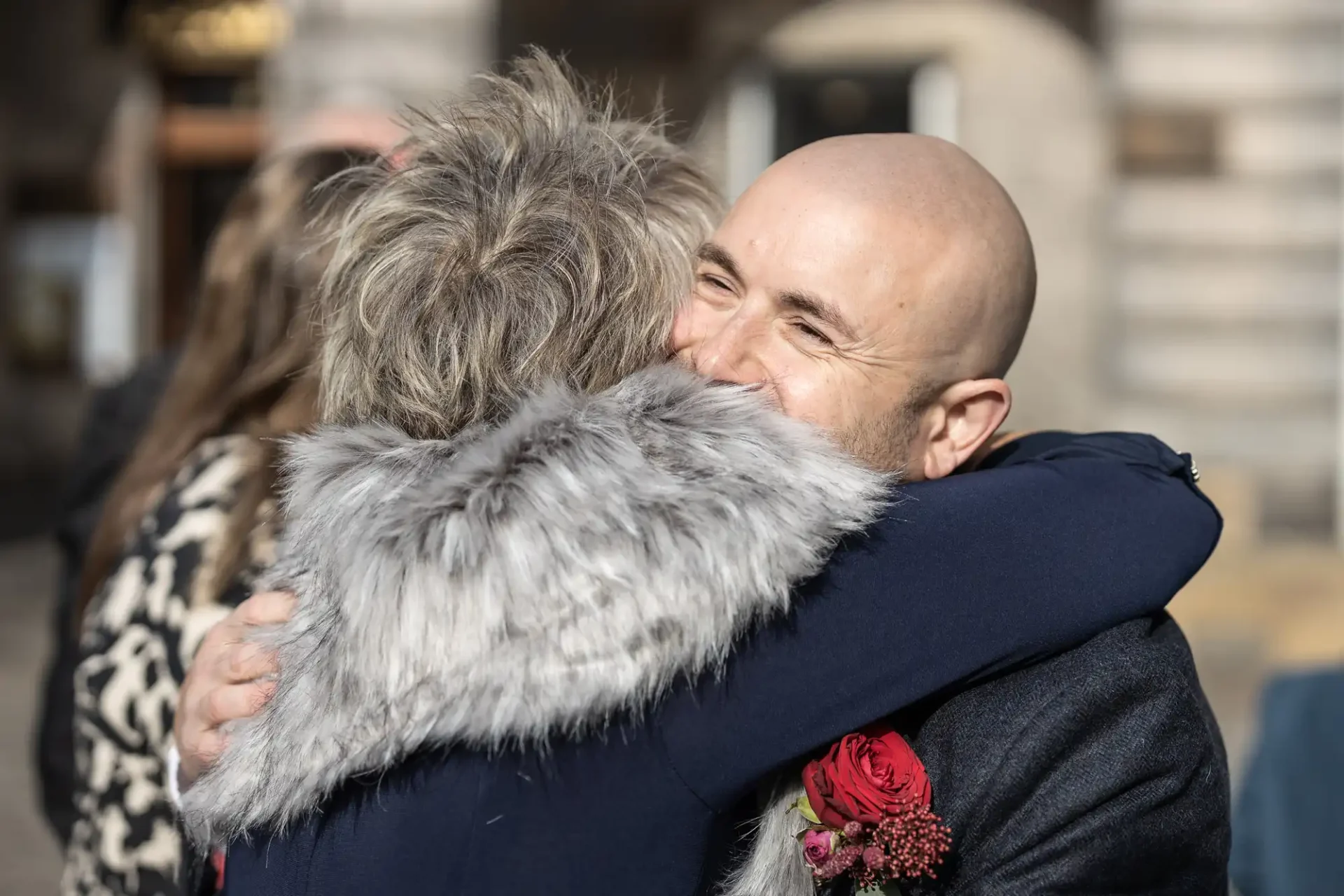 Two people hug outdoors, one with short hair and a gray fur-trimmed coat, the other bald with a red flower on their jacket.