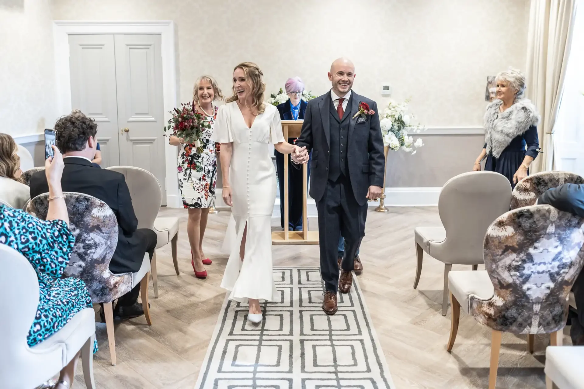 A couple in wedding attire walk down an aisle smiling. Guests seated on both sides, some taking photos. A person at a lectern in the background.