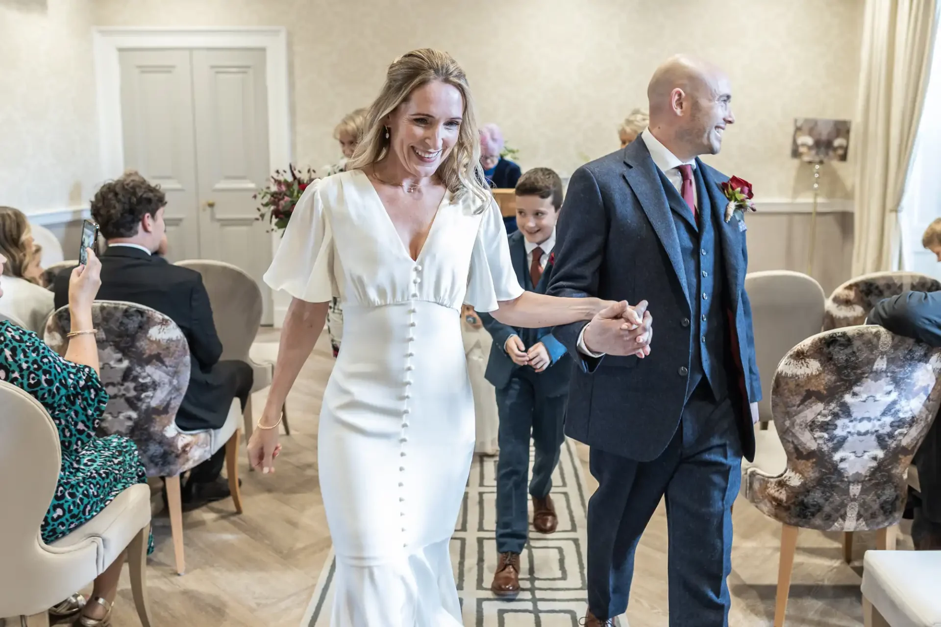 A smiling bride in a white dress and a groom in a blue suit walk hand in hand down an aisle in a small, decorated room with seated guests.