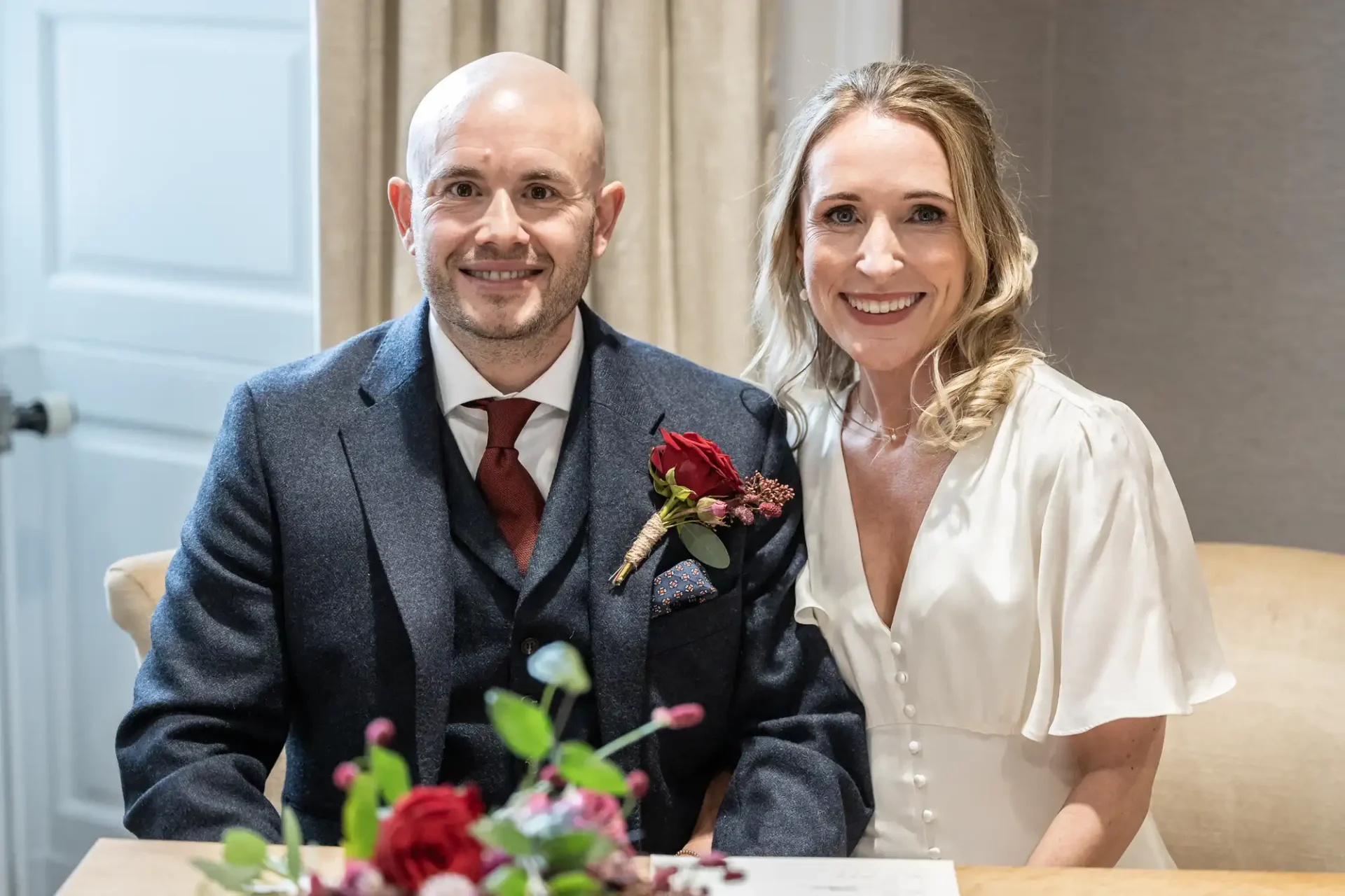 A couple sits at a table, smiling. The man wears a dark suit with a red tie and boutonniere, while the woman is in a white dress. A floral arrangement is visible in the foreground.