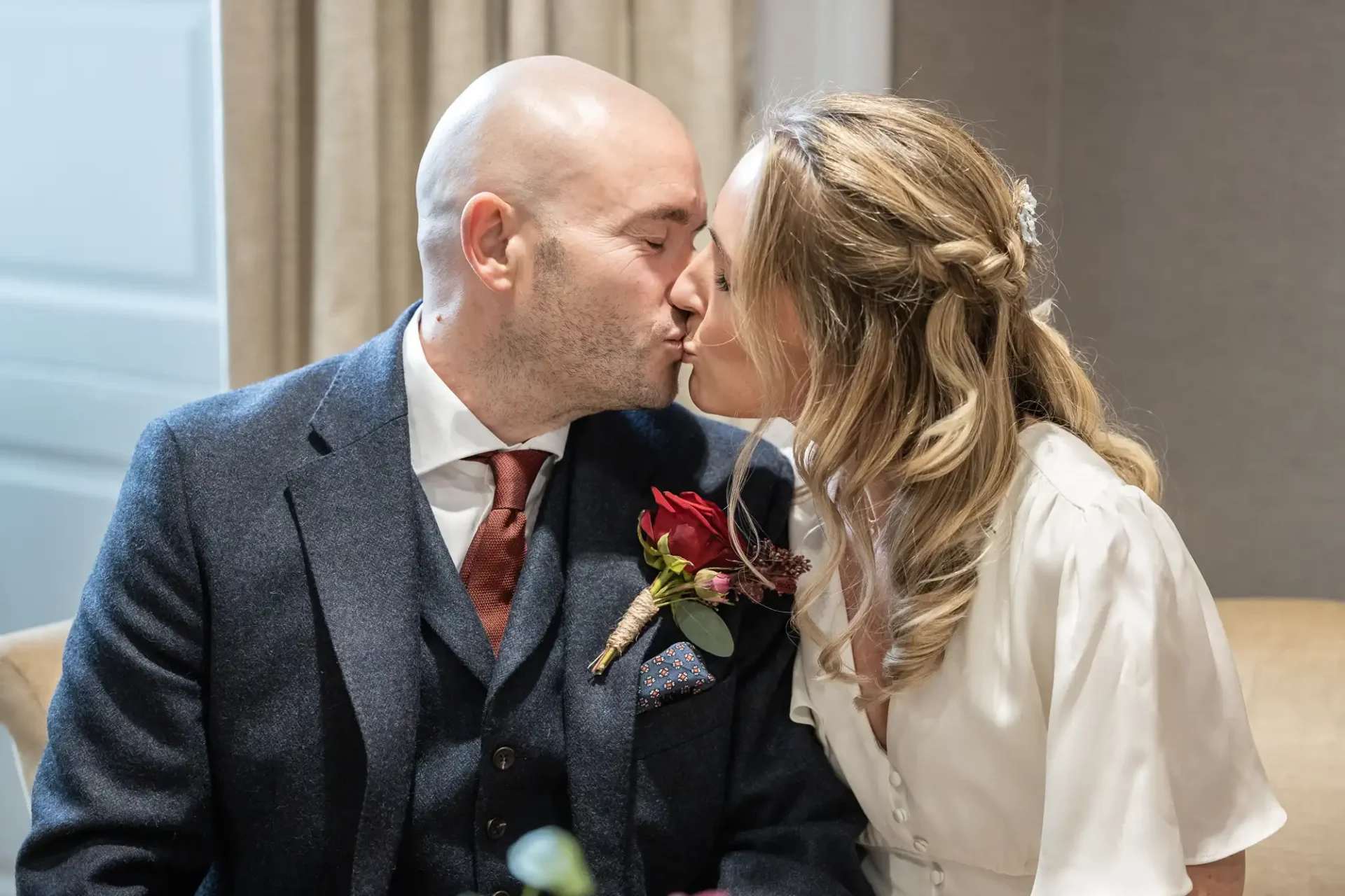 A couple in formal attire shares a kiss; the man wears a dark suit with a red tie and boutonniere, and the woman wears a white dress with wavy hair.