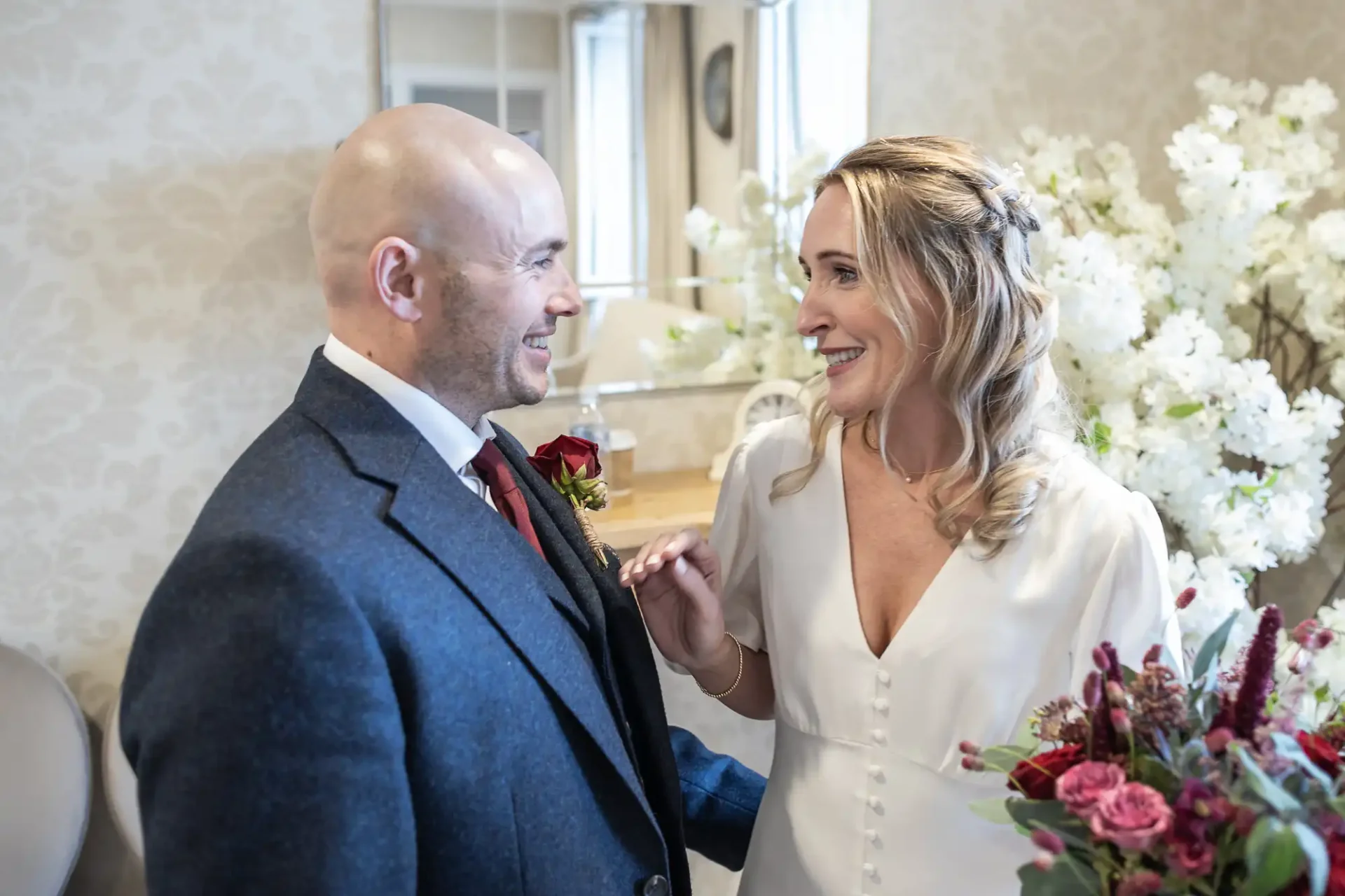 A couple stands together, smiling at each other. The woman holds a bouquet of flowers, and both are dressed in formal attire, with the man in a suit and the woman in a white dress.