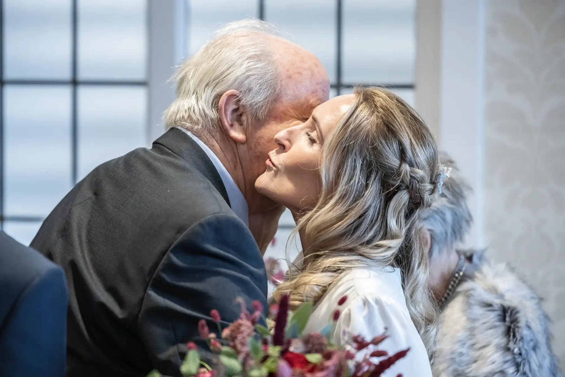 An elderly man and a woman embrace in a warm gesture. She holds a bouquet of red and pink flowers.