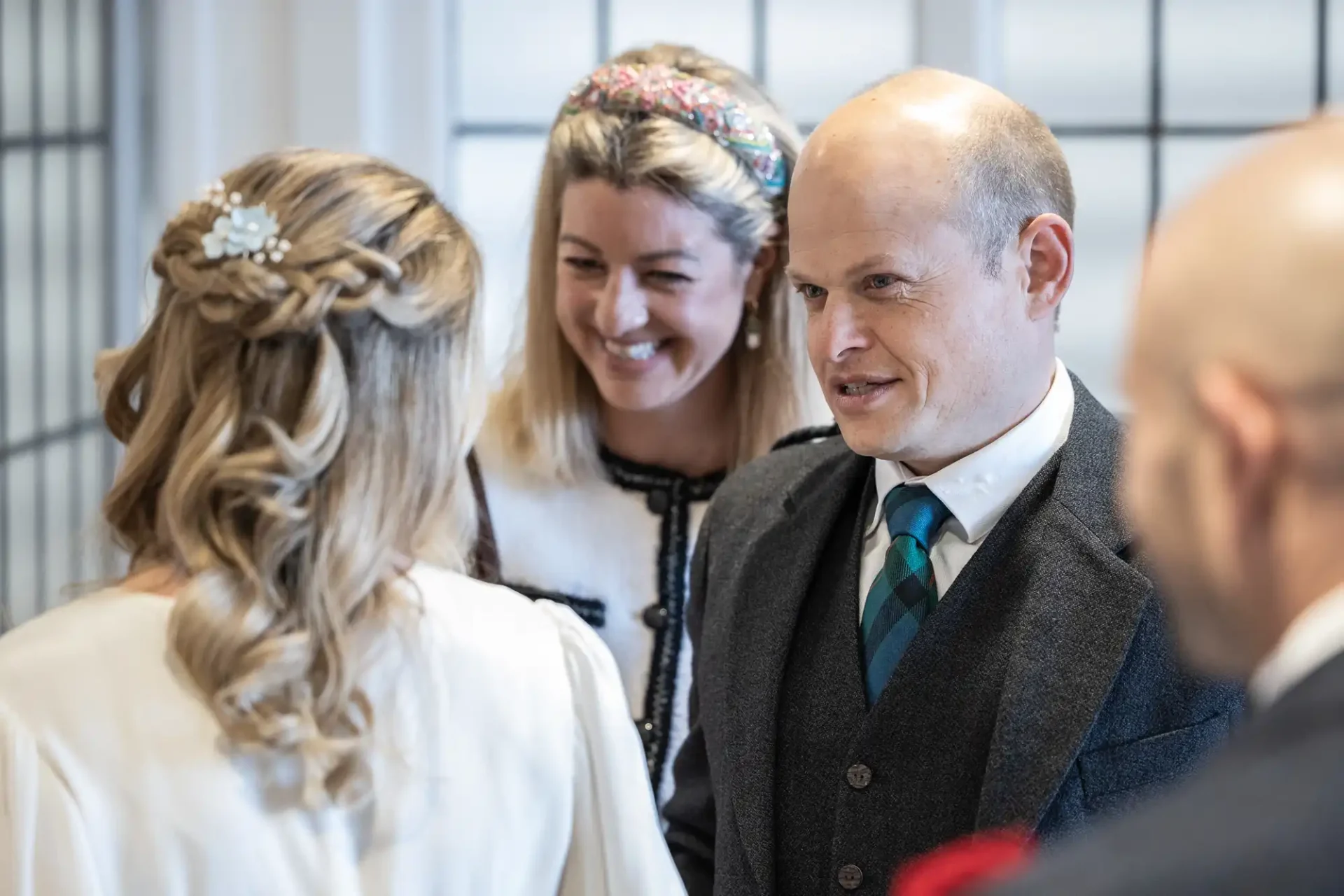 A woman and two men chat in a bright room. The woman has blonde hair and a patterned headband, one man has a bald head, and the other is partially visible. The room has large, grid-like windows.