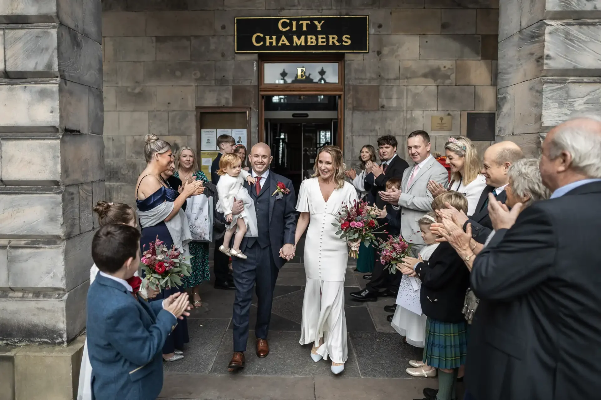 A newlywed couple exits a building marked "City Chambers," holding hands and smiling, surrounded by a group of people clapping and celebrating.