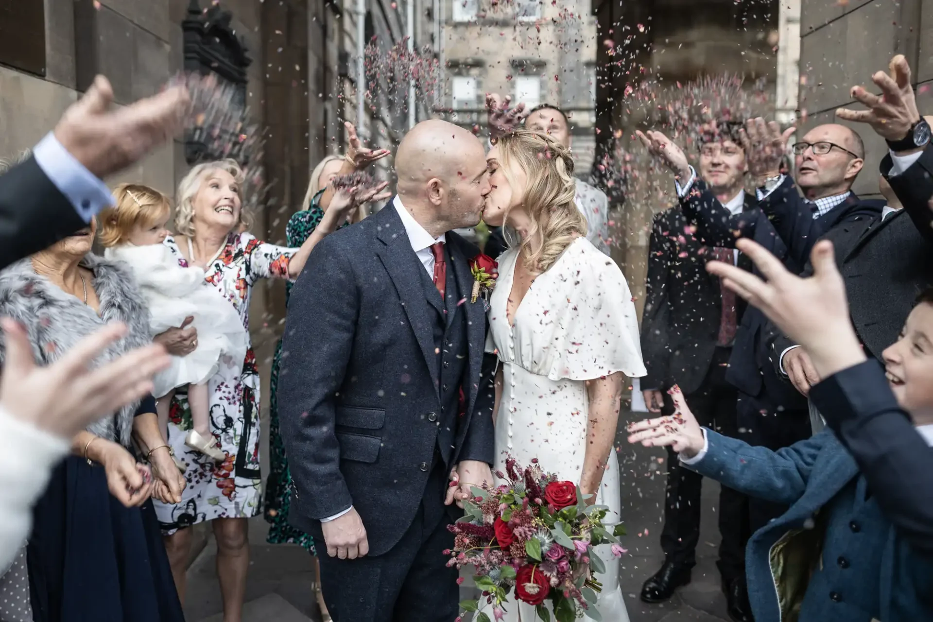 A couple kisses while confetti is thrown around them. They are surrounded by people celebrating. The bride holds a bouquet of red and green flowers.