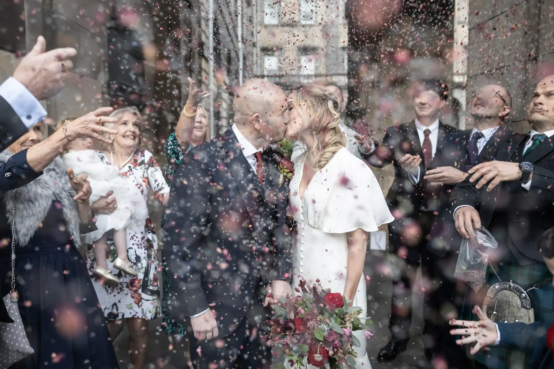 Bride and groom kiss as guests throw confetti; the bride holds a bouquet of red roses.