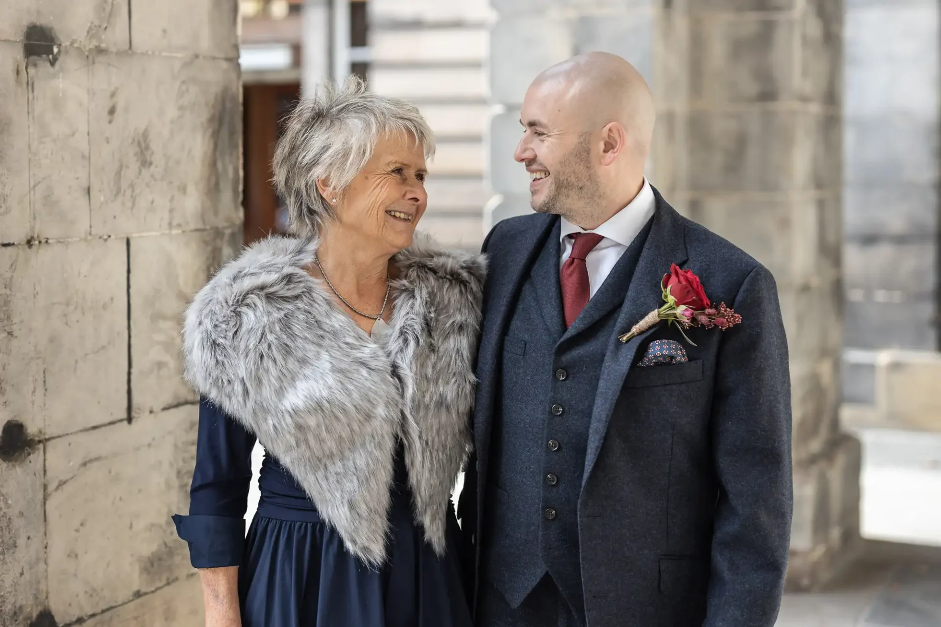 An older woman and a bald man in formal attire smile at each other, standing under a stone archway.